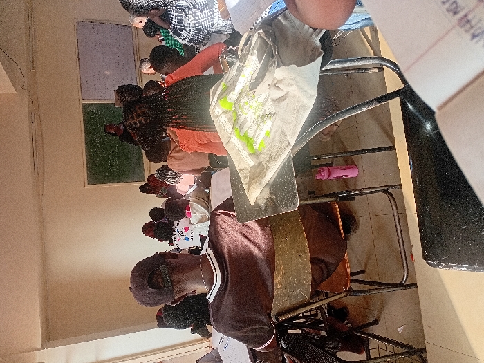 The image displays an indoor classroom in Karingani ward, Kenya, filled with approximately fifteen to twenty individuals, mostly students, attending an instructional session. In the foreground, facing away from the viewer, a male student with a dark baseball cap and a brown t-shirt is seated on a metal-framed chair with brown upholstery. To his immediate right, a female student with braided hair, wearing an orange top, sits at a desk that holds a light-colored item and several green writing implements. A pink water bottle is on the floor beneath a desk. Rows of students occupy similar metal-framed desks and chairs, facing a front wall where a female figure, presumed to be the lecturer, stands between a dark green chalkboard and a white board, both containing written information. Natural light enters the room from the left, illuminating parts of the scene. The walls are light-colored, and the floor is tiled.