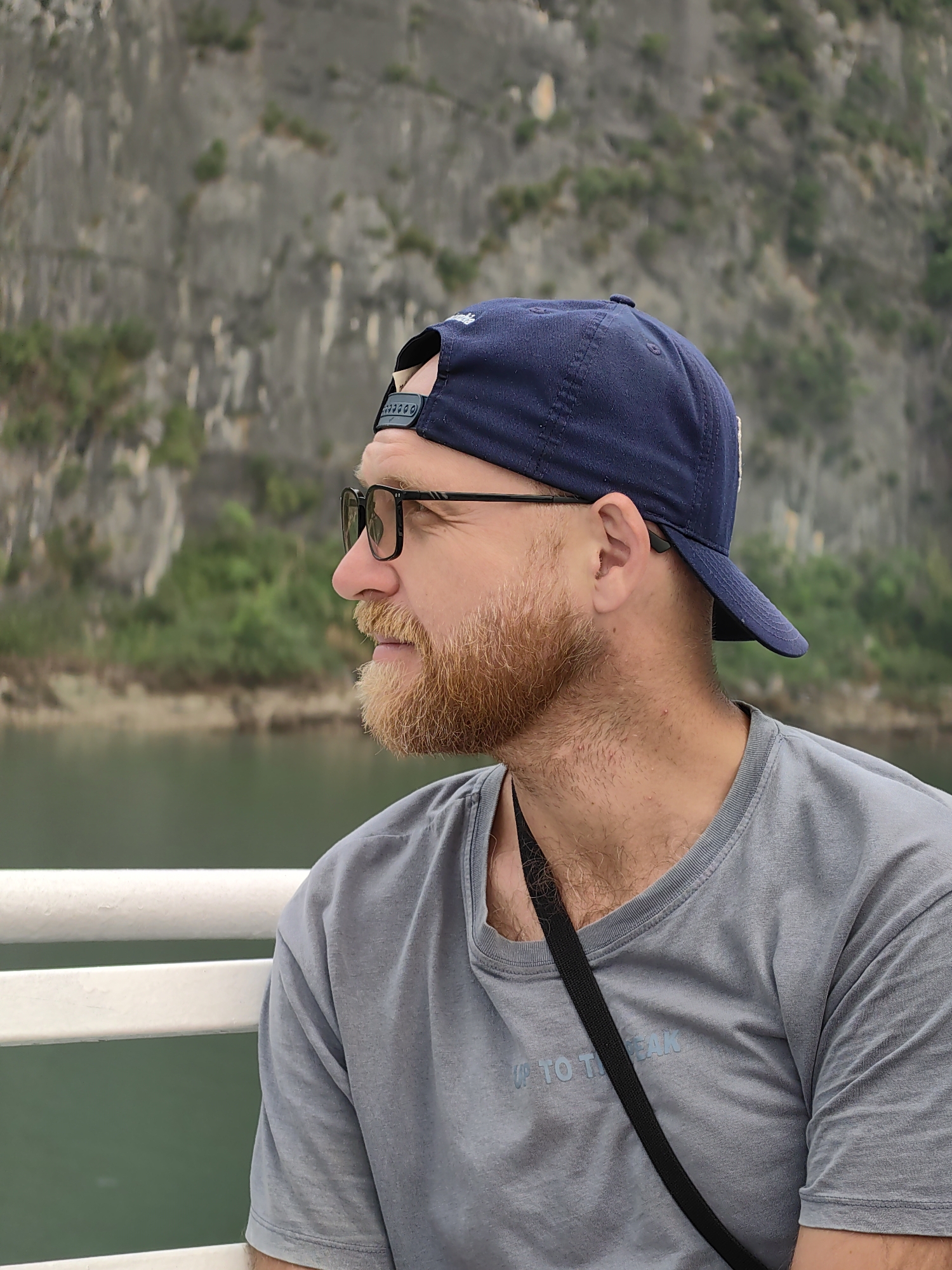 A man with a red beard and wearing glasses is seated on a boat, gazing to the left. He is wearing a blue baseball cap and a gray t-shirt. A black strap is draped across his chest. The background shows a body of water with a rocky cliff face covered in greenery. The location is likely Bai Chay Ward, Vietnam, due to the distinctive limestone formations.