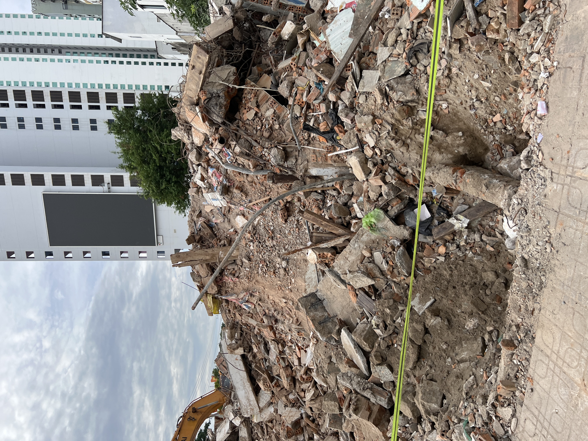 The image shows a large pile of rubble from a demolished building in Nha Trang, Vietnam. The rubble consists of broken concrete, bricks, metal rebar, and other debris. In the background, there are two tall, modern buildings, one of which has a large, dark screen on its facade. An excavator arm is visible on the left side of the rubble pile, suggesting ongoing demolition or clearing work. A neon yellow caution tape stretches across the foreground, indicating a restricted area. The ground in the foreground is paved with concrete tiles. The sky is overcast with clouds.