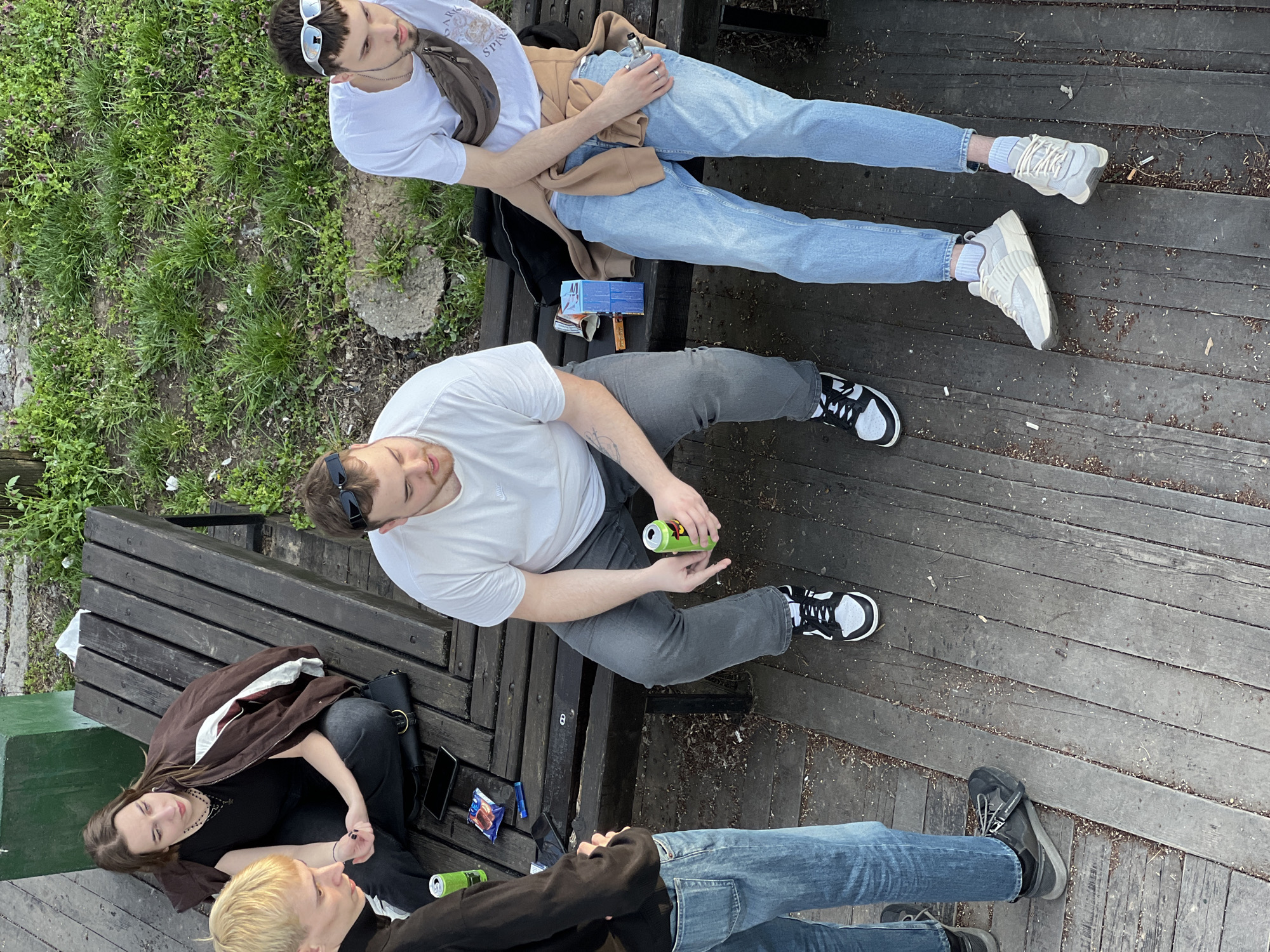 The image shows four young adults sitting on a wooden bench. One man is in a white t-shirt and light blue jeans. Another man is in a white t-shirt and gray pants, holding a green can. A woman in a black top and a brown jacket sits with a man in a black jacket. There are several objects on the bench including phones, a black purse, a vape, a box, and several green cans. The location appears to be an outdoor area. The location is Belgrade, Serbia.