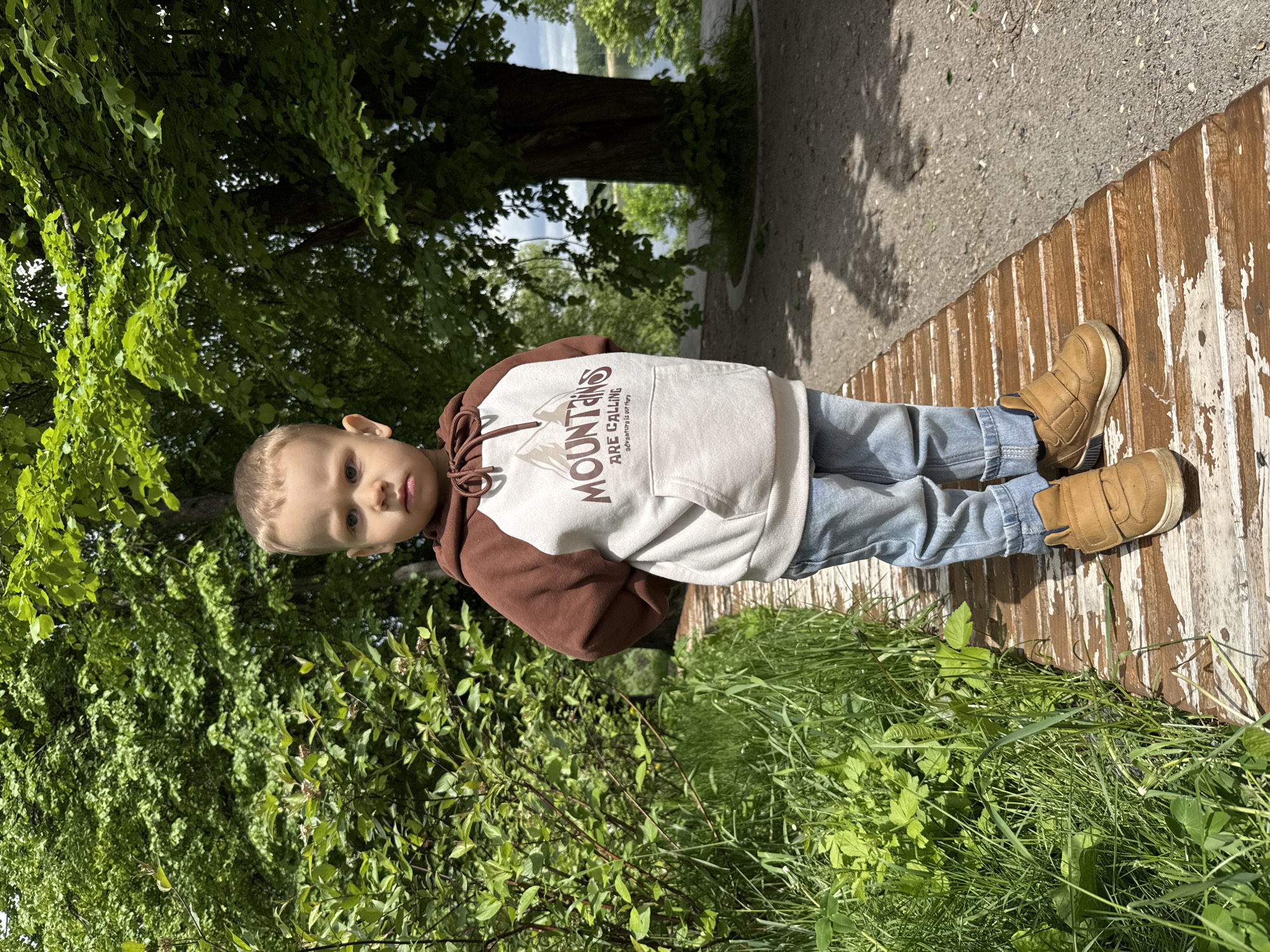 A young child stands on a weathered wooden structure, likely a walkway or bench, in an outdoor setting. The child wears a brown and white hoodie with "MOUNTAINS ARE CALLING" printed on it, blue jeans, and tan shoes. Lush green vegetation surrounds the walkway, and a tree is visible in the background. In the distance, a body of water is visible. The location appears to be in Rusanovo, Russia.