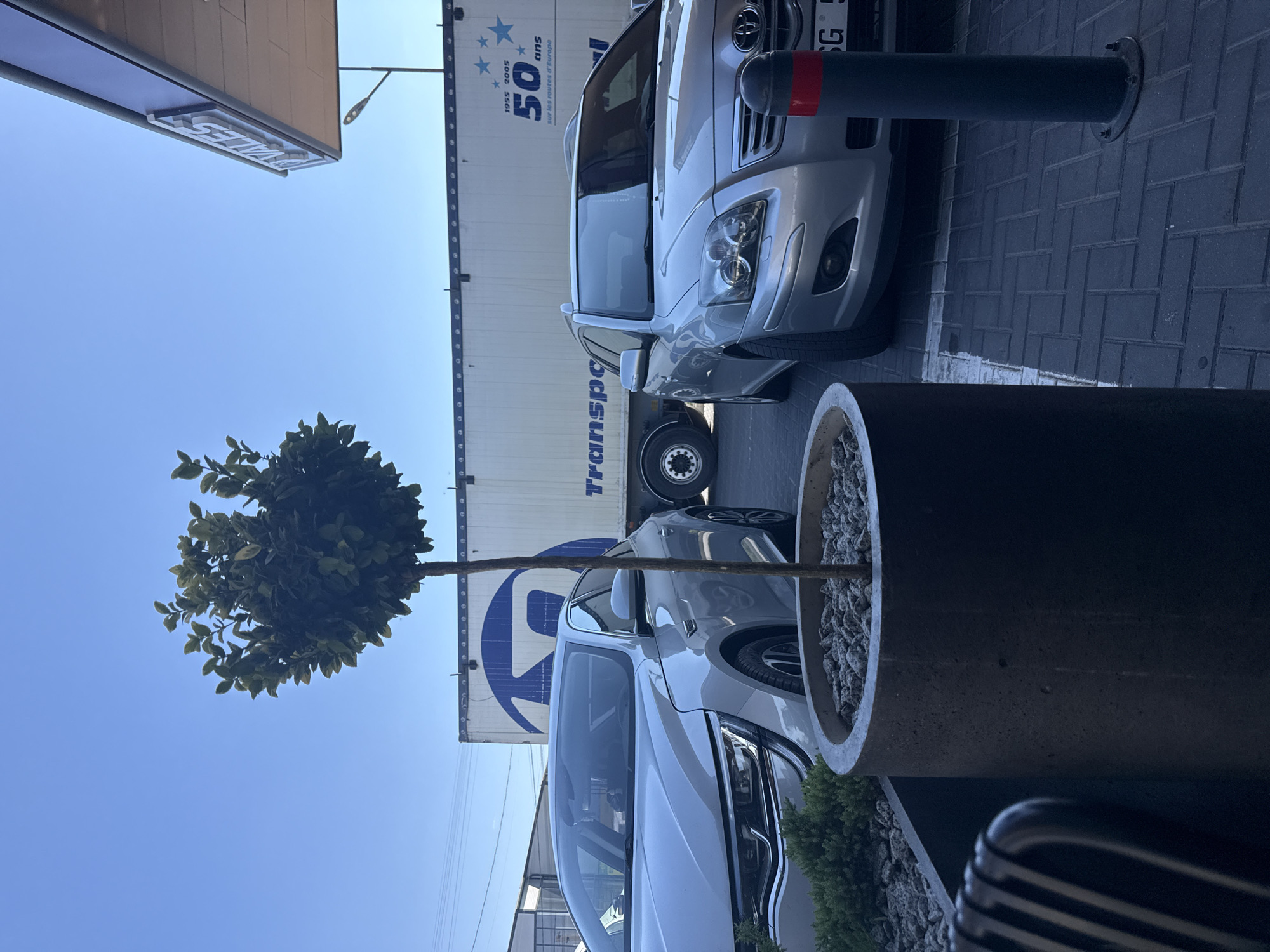 The image depicts an outdoor scene, possibly a parking lot or a service area, under a clear blue sky. In the foreground, a dark gray, cylindrical planter contains a small, leafy tree. To the left of the planter is a silver sedan, and to its right is a silver station wagon. Behind the cars, a large white semi-trailer truck is parked, displaying a large blue circular logo on its side with white lettering that appears to read "TRANSPO". Further to the right, a sign with white lettering that says "VALES" is visible, attached to the overhang of a building that has a light fixture extending from it. The ground is paved with dark gray interlocking bricks. A black bollard with a red stripe is situated in front of the station wagon. In the background, beyond the truck, a building with white siding and windows is visible. A portion of a metal fence or railing is also present in the background. The overall scene suggests a commercial or transportation-related location. Based on the provided information, this location is identified as being in the city of Comrat, Moldova.