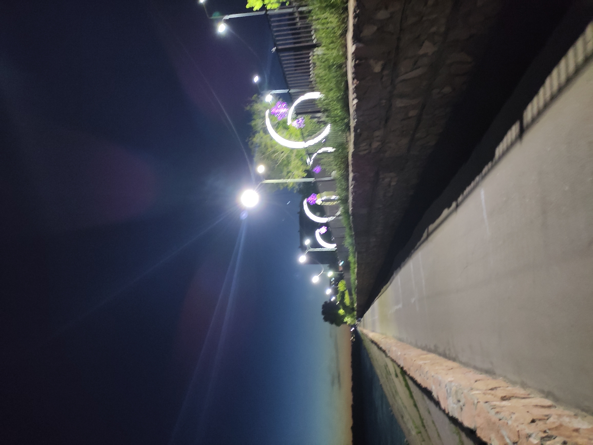 This is a low-angle, vertical-format night photograph of a city scene along a river embankment. On the left, a dark sky transitions from a lighter horizon toward the bottom. A line of lampposts with decorative crescent moons is positioned along a path. The walkway next to the river is framed by stone walls and appears to be deserted, though there are a few people or objects in the distance. The photo appears to be taken on the Pavlodar embankment in Kazakhstan.