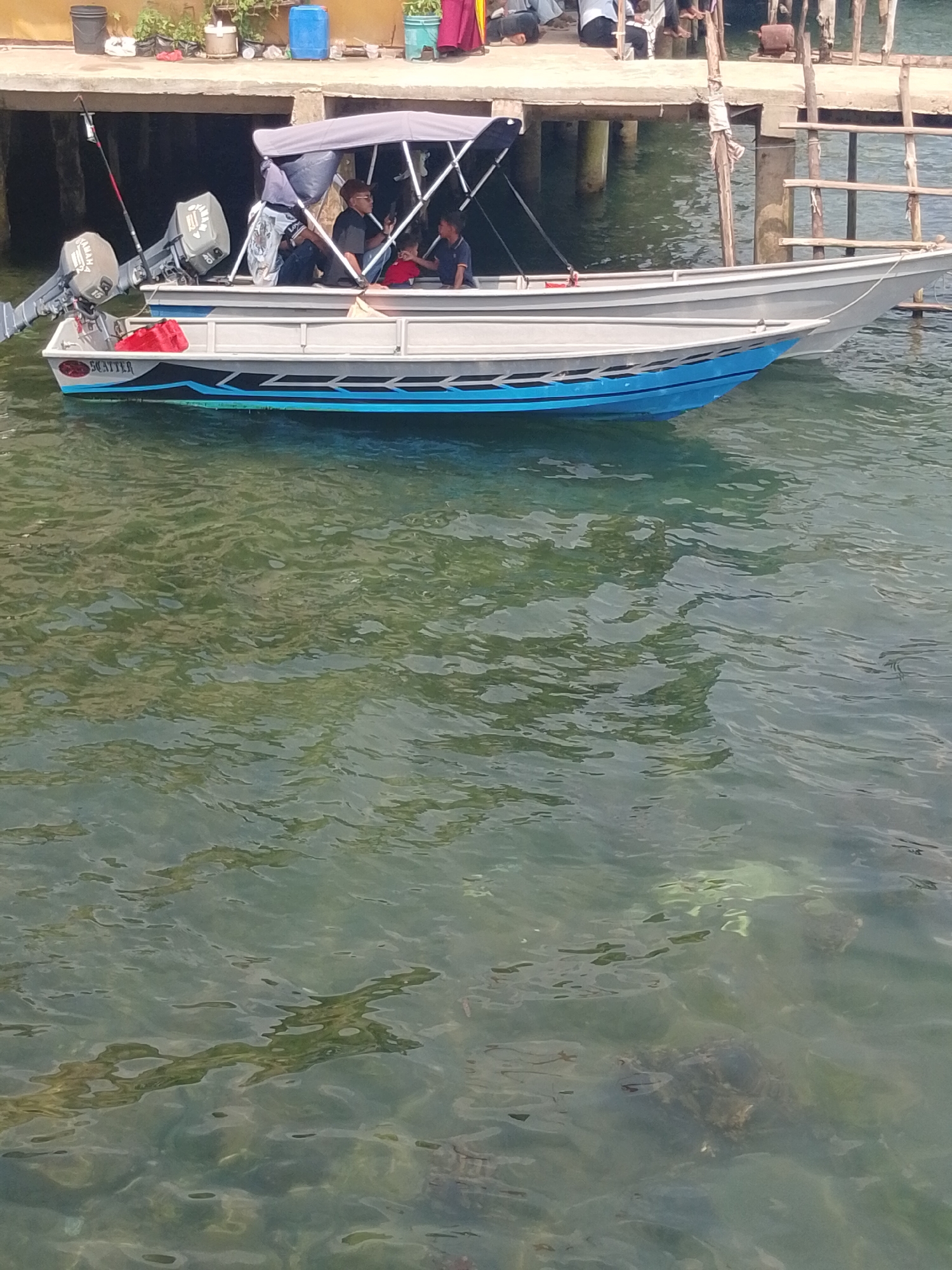 In Batam, Indonesia, a boat with a canopy is docked near a pier. Two men are seated in the boat. One man is wearing a dark shirt and sunglasses, and the other is wearing a blue shirt. A red container is visible in the boat, along with two outboard motors. The water is clear and the bottom is visible. The pier is constructed of wood and has some debris on it. The scene appears to be a typical waterfront view in a tropical location.