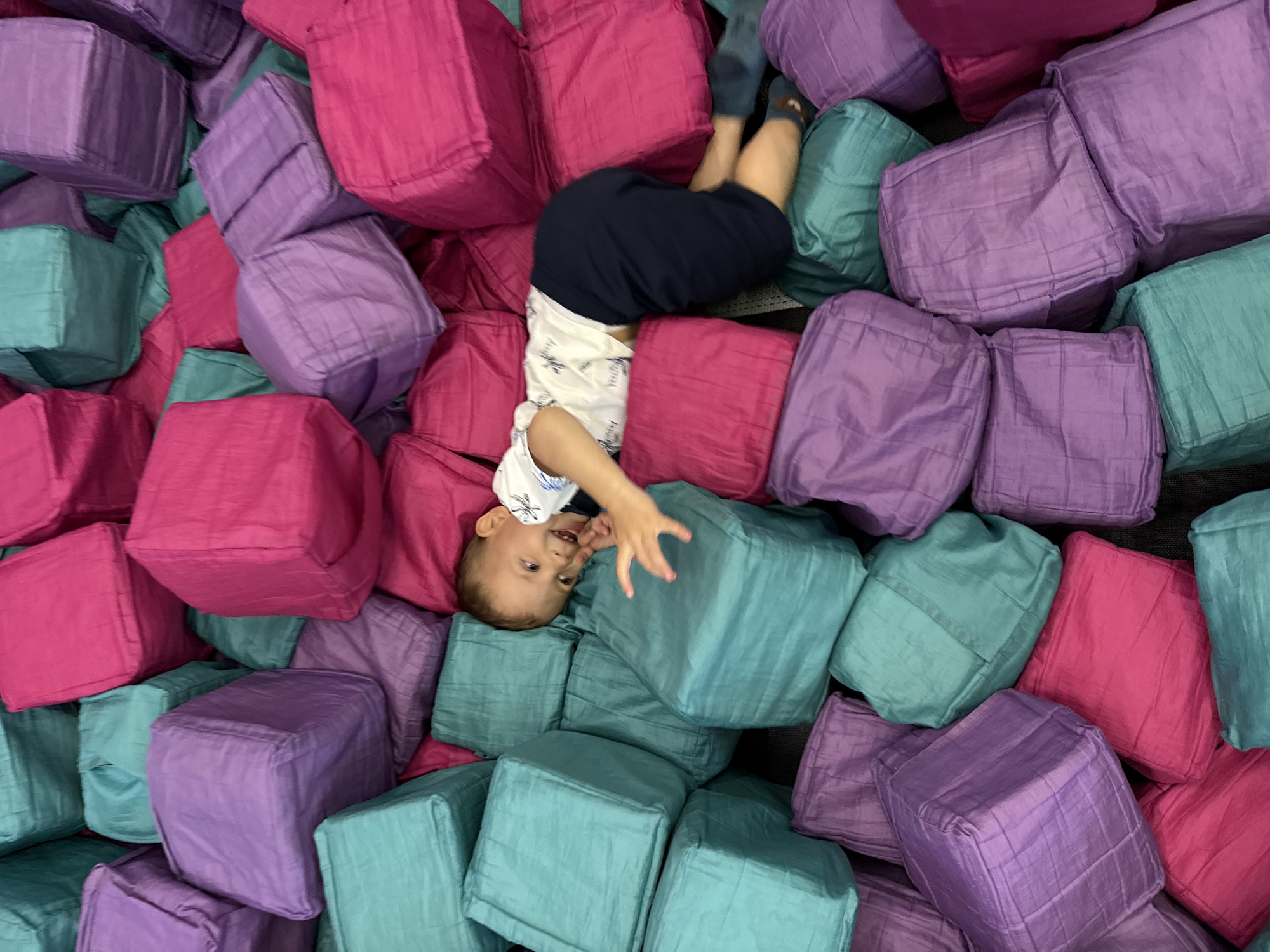 A young boy lies in a pile of colorful, soft cubes in a foam pit. The cubes are a mix of vibrant pink, teal, and purple. The boy, wearing a white shirt with a black print and dark blue shorts, is lying on his side, facing right and looking towards the viewer with a wide smile. His left arm is extended, and his hand is reaching out towards the cubes. The background is entirely filled with these cubes, creating a soft and playful environment. The scene appears to be indoors, likely in a children's play area or an indoor playground. The image is taken from a high angle, looking down into the foam pit. There is no indication of the specific location within the city of Zhukovo, Russia, in the provided image or context.