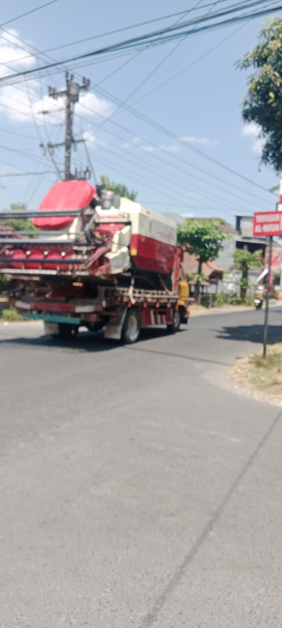 An outdoor, eye-level, medium shot captures a harvester being transported on a truck driving down a paved road in Maguwoharjo, Indonesia. The truck, predominantly red and white with a yellow cab, carries a large, agricultural harvesting machine on its flatbed. The harvester's red canopy and red and silver machinery are visible. Power lines stretch across the bright blue sky, crisscrossed by a utility pole with electrical equipment. Lush green trees frame the scene on the right side, and a red and white signpost with Indonesian text is visible in the background. The road is gray asphalt, with a slight curve to the right, and some dry grass and dirt line the edge of the road. The image is slightly blurred, indicating motion.
