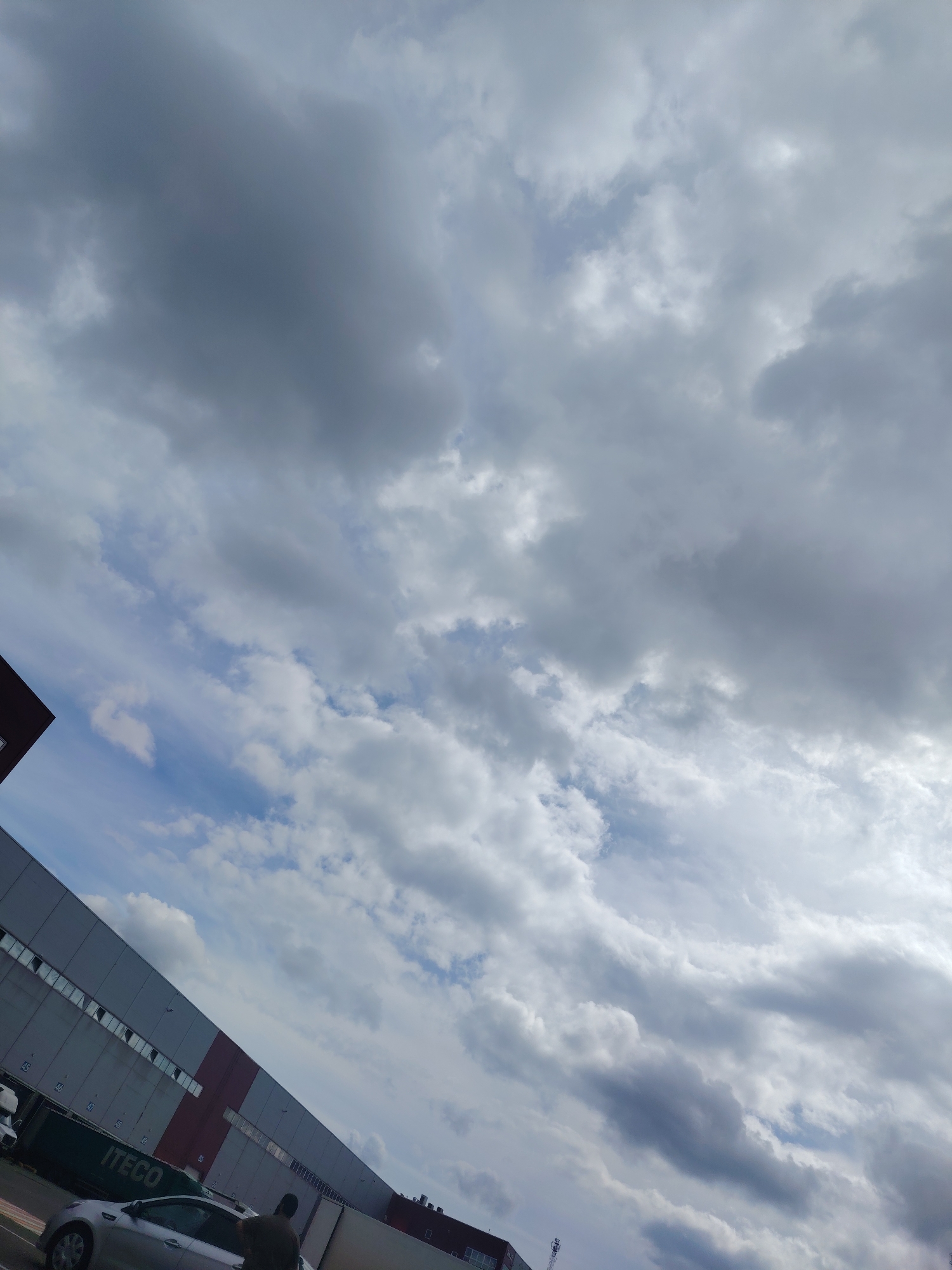 The image, taken in Khorugvino, Russia, depicts a vast, overcast sky filled with a mix of dark grey and white clouds. The sky occupies most of the frame, with a sliver of a building complex visible in the lower left and bottom of the image. The building appears to be a large industrial or warehouse structure with a red and grey facade. Several trucks and cars are parked in front of the building. One truck has "ITECO" written on its side. A silver car is parked near the foreground, and a person can be seen standing near it, partially obscured by the car. The overall scene suggests a daytime setting under a cloudy sky, possibly in an industrial or commercial area of the city.