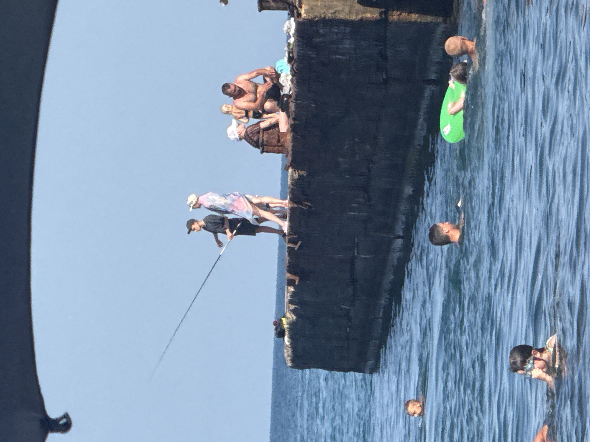 In Partenit, Ukraine, on a sunny day, several people are gathered on a concrete pier and in the adjacent water. A young male stands on the pier, fishing with a long rod. Beside him, a young female in a light-colored cover-up stands, and behind them, a man and a child in swimwear are seated. Further down the pier, other individuals are visible. In the water, several people are swimming, with one person using a green inflatable ring. The sea is calm with small waves, and the sky is clear and blue. The pier appears old and weathered.