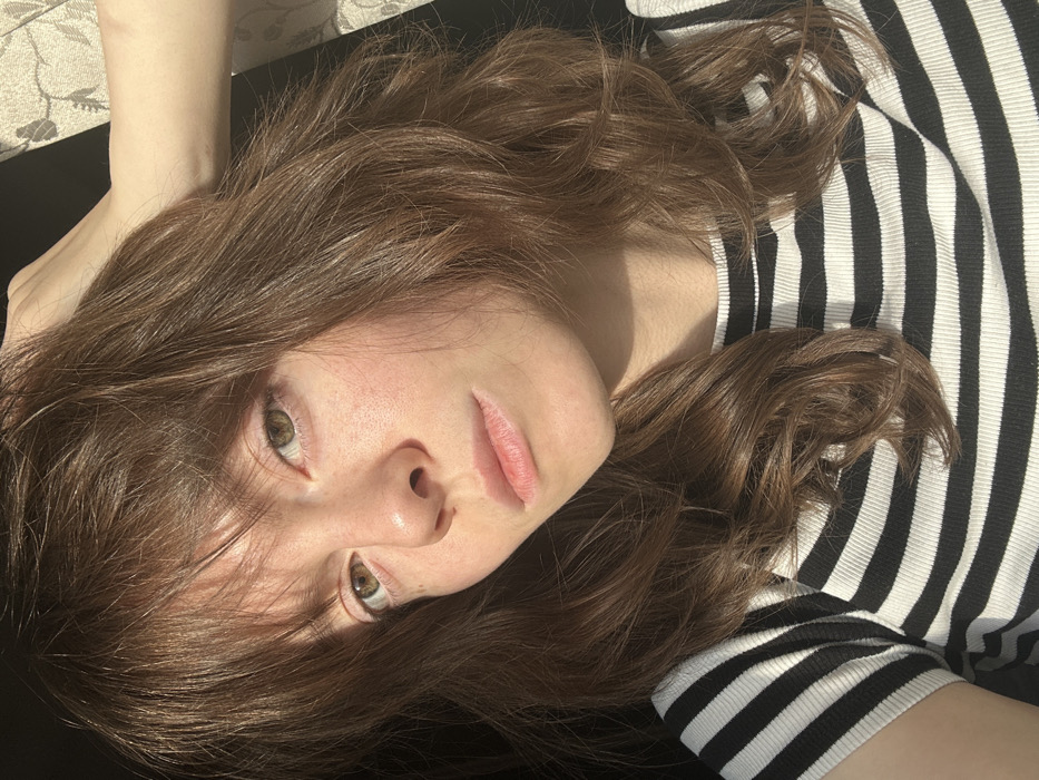A young adult female is depicted in a head-and-shoulders portrait. She has medium-length, wavy brown hair with bangs, hazel eyes, and a fair complexion with visible freckles across her nose and cheeks. Her lips are closed in a neutral expression. She wears a black and white horizontally striped, ribbed short-sleeved top. Her right hand is raised, with fingers resting in her hair near the crown of her head, appearing to adjust it. Strong natural light illuminates the right side of her face and hair, casting the left side into shadow. The background consists of a dark, indistinct area to her left and a partially visible light-colored curtain with a subtle pattern to her right, indicating an indoor setting near a window. The image contains no identifiable landmarks or specific features to determine a precise geographical location, such as Moscow, Russia.