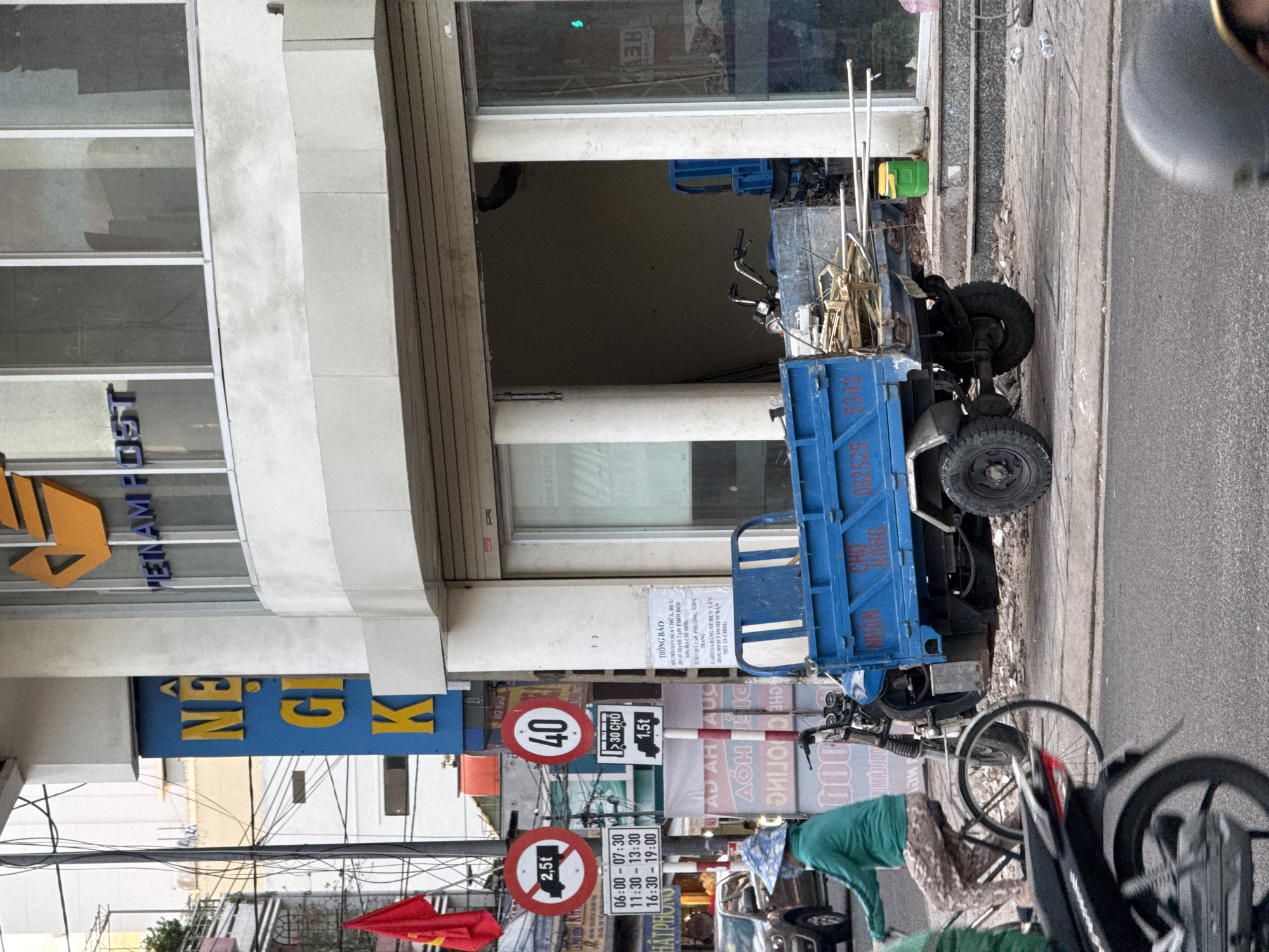 The image captures a street scene in Nha Trang, Vietnam, with a focus on a blue three-wheeled vehicle laden with construction materials. The vehicle is parked on a sidewalk adjacent to a building with large windows and signage for "VIETNAM POST." Several traffic signs are visible, including one indicating a weight limit of 2.5 tons and another displaying a speed limit of 40. A person wearing a green jacket and a patterned skirt is riding a bicycle in the foreground, partially obscuring a motorcycle. The ground is paved, with debris and construction materials scattered around. The overall impression is a busy urban environment, characteristic of a commercial district in Vietnam.