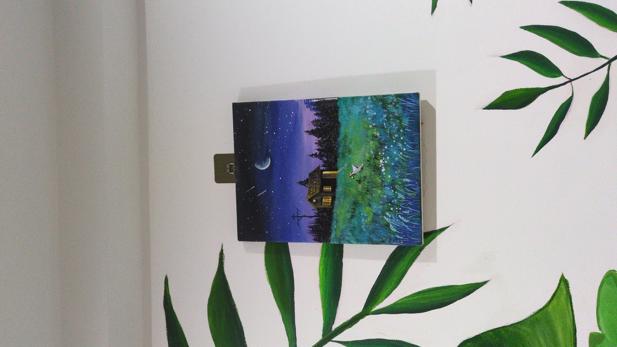 A square painting hangs on a white wall, held by a small, dark-colored clip. The painting depicts a landscape scene with a dark blue, star-filled night sky featuring a crescent moon and shooting stars. Below the sky, a line of dark green trees borders a field of green and blue grass with small white flowers. In the center of the painting, a house with lighted windows stands. A small white bird flies above the grass. The wall also has painted green leaves, resembling a plant. There is no available information about the image being located in the city of Phan Rang – Tháp Chàm, Vietnam.