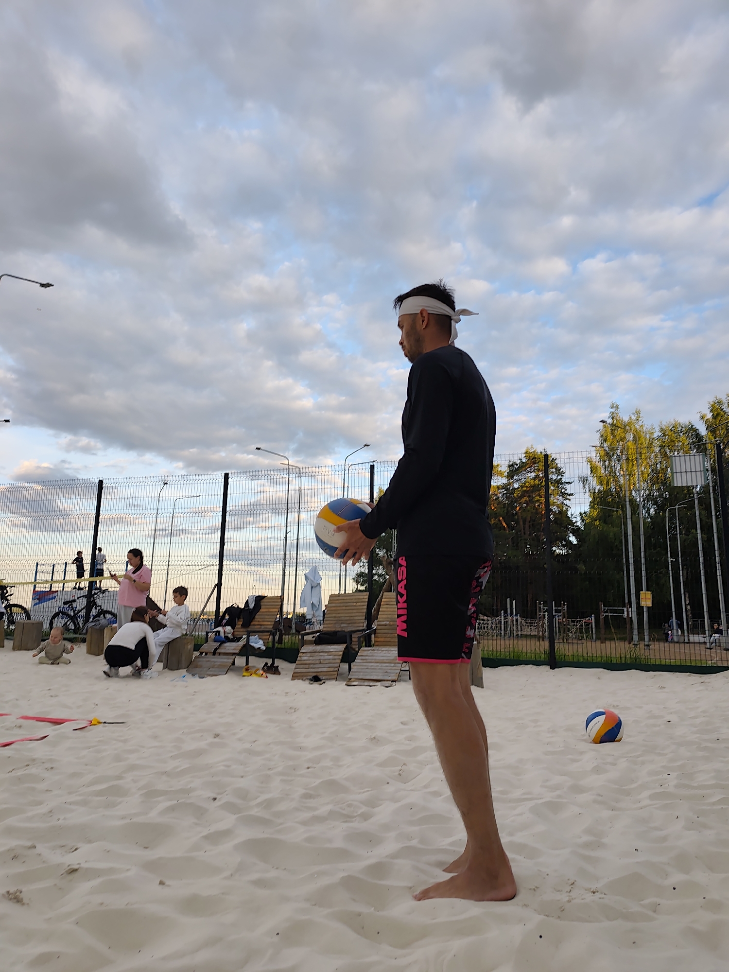 A man is playing volleyball on a sandy court. He is wearing a black long-sleeved shirt and black shorts with pink trim. He has a white headband on. He is holding a volleyball in his left hand. In the background, there is a fence, trees, and a cloudy sky. Other people can be seen in the background, including children and adults. A baby is sitting on the sand in the foreground. A volleyball is also visible on the sand in the foreground, to the right of the man. The time of day appears to be late afternoon or early evening, given the lighting. The weather is cloudy. The text "MIKASA" is visible on the man's shorts.