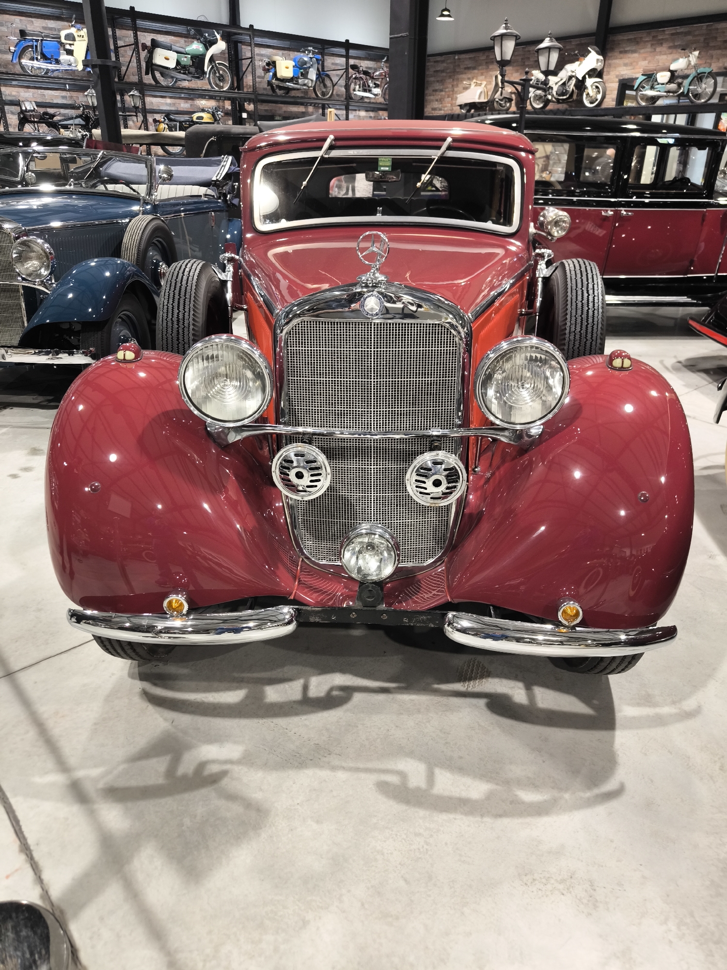 The image shows the front of a vintage red Mercedes-Benz car, possibly a 1930s model, prominently displayed in what appears to be a car museum or showroom. The car is detailed with chrome features on its grille, headlights, and bumper. Behind the Mercedes, other vintage cars and motorcycles are visible, arranged on shelves and on the floor, indicating a collection setting. The environment seems indoor with neutral grey flooring and adequate lighting. There is no visible evidence to confirm this is located in Kapatovo, Bulgaria.