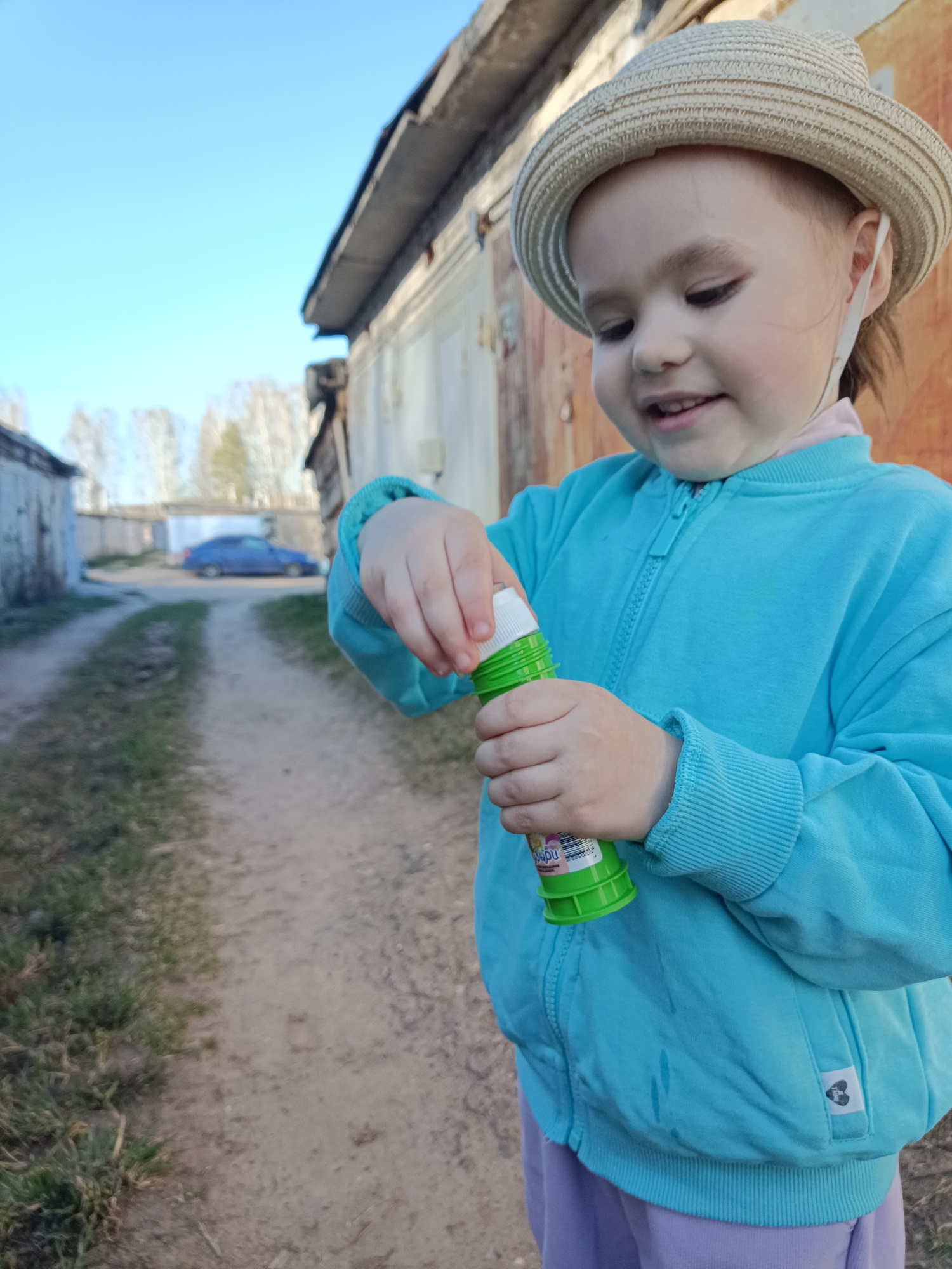 A young child in a straw hat and blue jacket stands on a dirt path near a row of garages. The child is opening a green bottle of bubble solution. The location is likely in the city of Chaikovsky, Russia, as per the instructions. A blue car is visible in the background on the path leading away from the child.