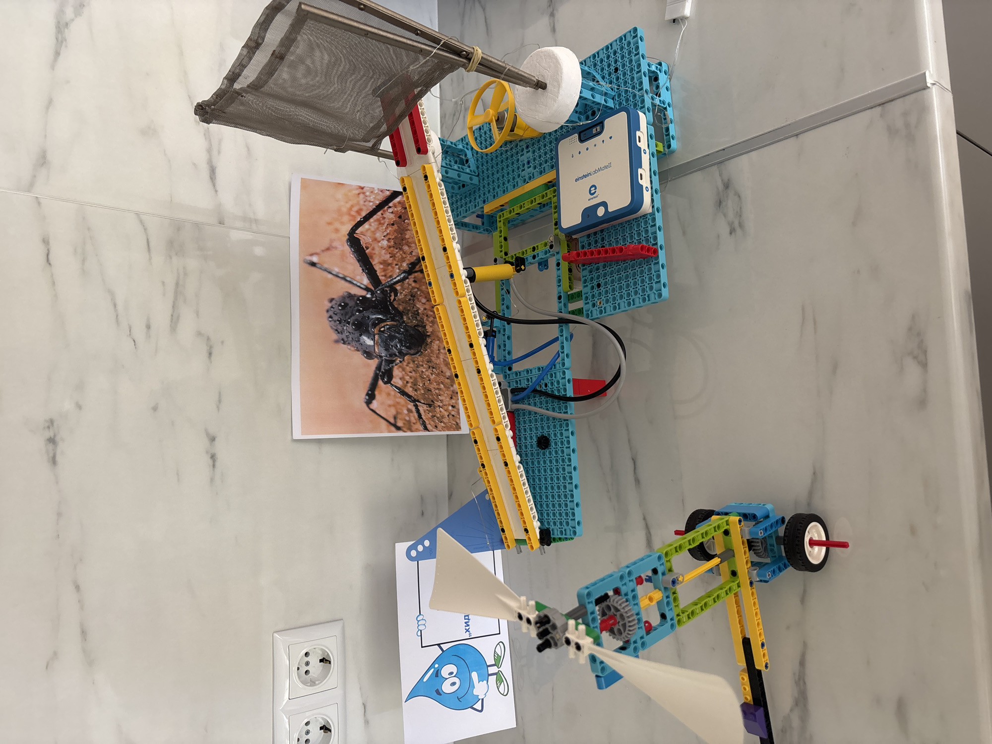 The image shows a homemade contraption attached to a white wall with a marble pattern, possibly in an educational or workshop setting. The contraption is constructed primarily from blue, yellow, green, red, and white Lego-like plastic building blocks and includes a mesh-covered frame, a white foam cylinder, a yellow wheel, a blue electronic device labeled "EinsteinLabMate II," and a small propeller with white blades. The building blocks are arranged to form a framework. A picture of a black beetle is attached near the top of the construction. Below that, a cartoon drawing of a smiling blue water droplet with the text "ХИД" (HID) written on a sign is visible. An electrical outlet with two sockets is visible in the lower left corner of the frame. There is no clear indication that the image was taken specifically in Zlatograd, Bulgaria.