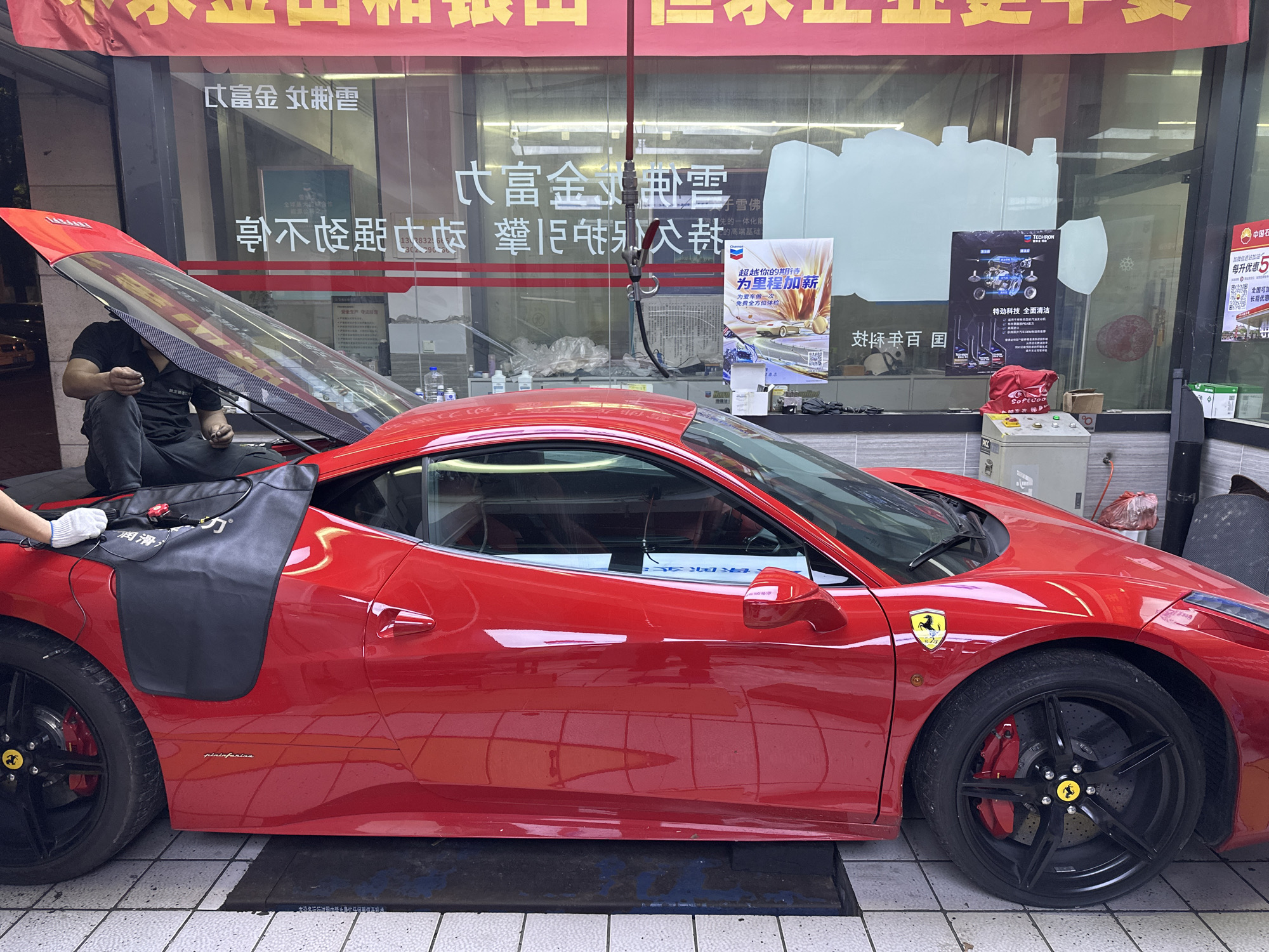 A bright red Ferrari sports car, likely a 458 model, is parked partially inside a car service or detailing shop in Chongqing High-tech Zone, China, during the daytime. The car's rear engine cover is open, revealing the engine bay, and a person in a black shirt and pants is sitting on the edge of the engine compartment, looking down at something in their hands, suggesting maintenance or detailing work. A black protective mat with "润滑" (Lubrication) partially visible is draped over the rear fender. Another person's foot, wearing white shoes, is seen near the rear of the vehicle.
The Ferrari features black multi-spoke wheels with red brake calipers and yellow Ferrari crests. The "pininfarina" script is visible on the side of the car, denoting its design heritage.
The background consists of a large glass storefront window reflecting its surroundings. Inside the shop, various bottles, cleaning supplies, and a red bag labeled "Softcoo" are visible on a counter. A hose/nozzle system hangs from the ceiling. Several promotional posters are displayed inside the window, including one for "TECHRON 特劲科技 全面清洁" (Techron special strength technology, comprehensive cleaning) and another with Chinese text "超越你的期待 为里程加新" (Exceed your expectations, add new mileage) and "为爱车做一次 免费全方位体检" (Give your beloved car a free comprehensive check-up). A banner above the shop window reads "富金 雲" (Rich Gold Cloud) and other Chinese characters. More Chinese text, such as "雪佛" (Chevrolet), "中国石" (China Petro), and promotional offers like "每升优惠5" (5 discount per liter), are also visible on the window.