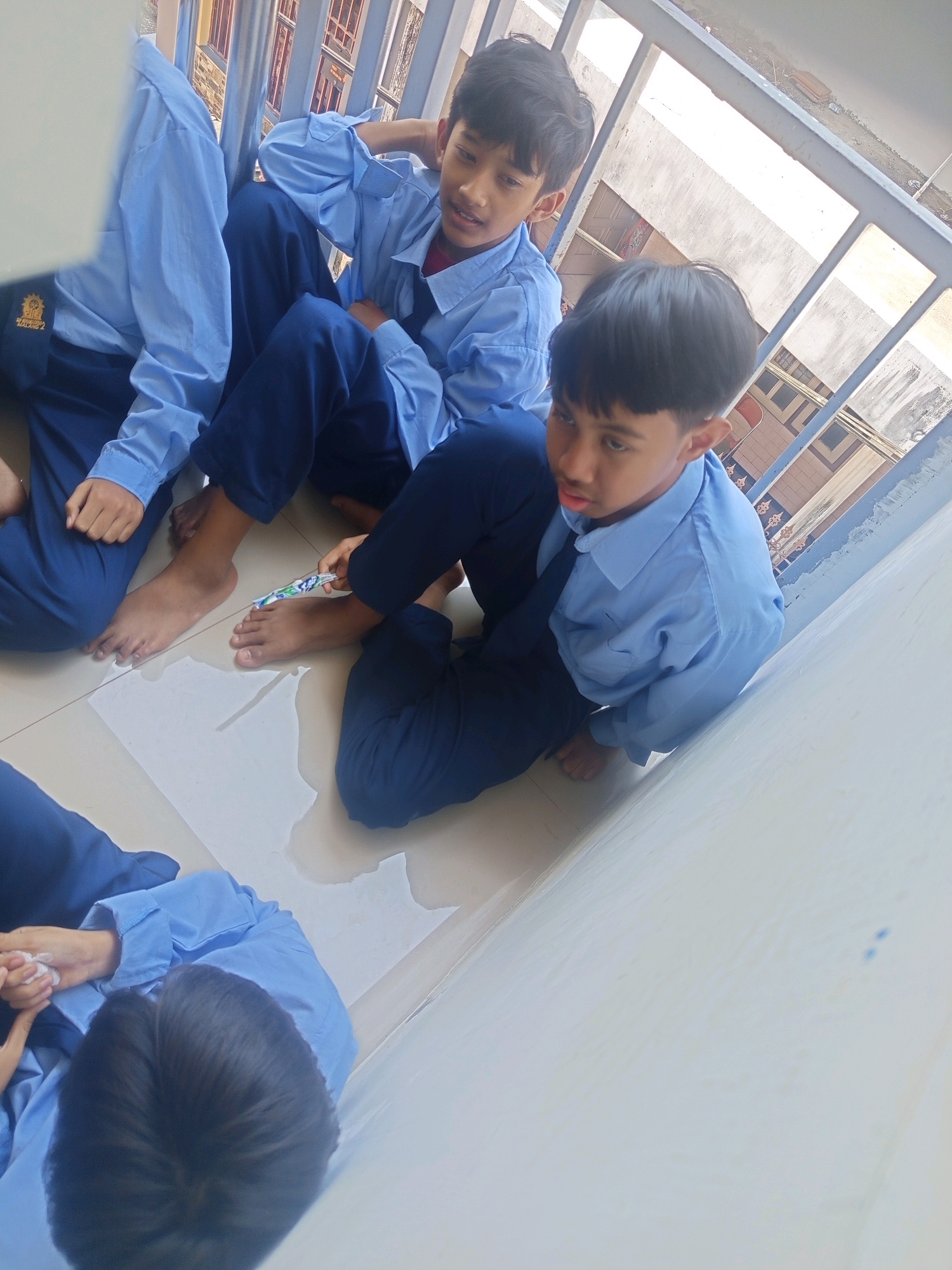 The image shows four young boys sitting on the floor, seemingly in a public space with a railing in the background. The boys are wearing blue shirts and dark blue pants, resembling school uniforms. One boy is holding a small object that appears to be a toothbrush with toothpaste. The environment suggests an outdoor or semi-outdoor setting, possibly a balcony or a walkway. The image is taken from a slightly elevated angle, looking down at the boys. The presence of a railing and a building structure in the background indicates an urban setting. The floor appears to be tiled. There is some water or liquid spilled on the floor. The overall scene depicts a casual moment among young students. The text on the logo on the left boy's shirt is not legible but may provide a clue to their school or affiliation. There is no explicit indication of the city of Malang, Indonesia within the visual elements of the image.