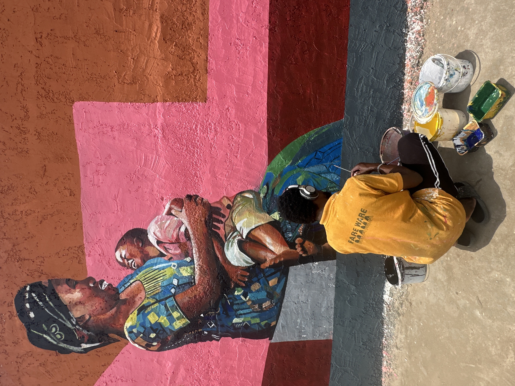 In the city of Badagry, Nigeria, a mural depicts a woman in a colorful headscarf and patterned dress holding a laughing child. The mural spans a textured wall painted in sections of orange, pink, and red. In the foreground, an individual wearing a yellow t-shirt with the text "FARE WARE MAMA" and headphones crouches on the ground, actively painting the mural. The painter is surrounded by several buckets of paint in various colors, including white, yellow, blue, and green, along with paint trays and brushes. The ground is paved with concrete.