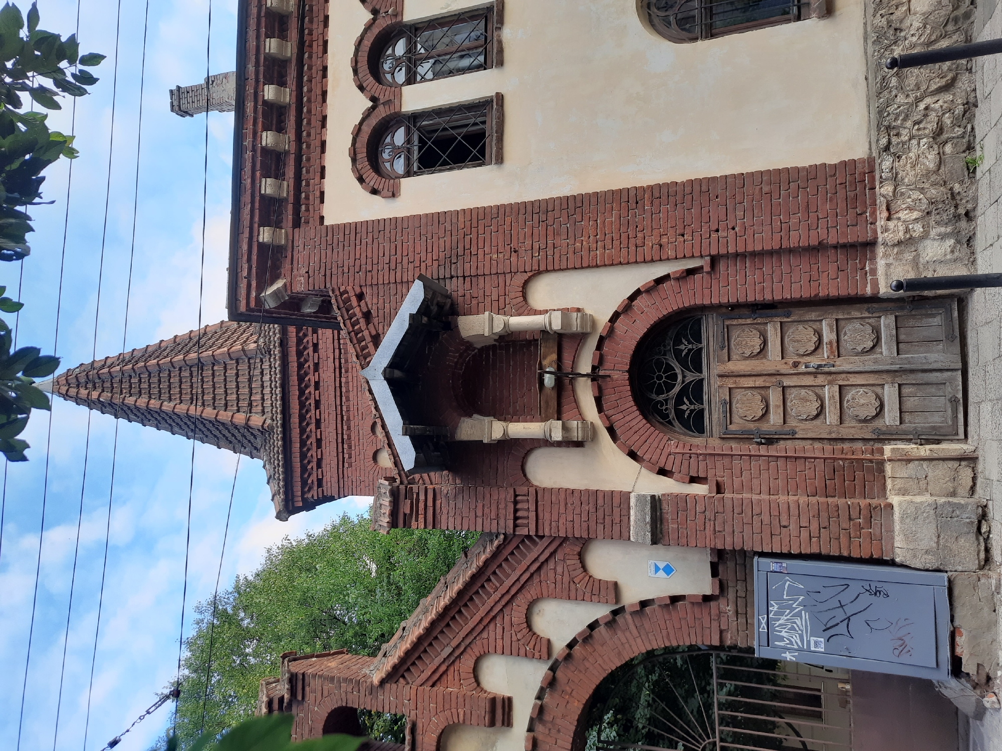 A brick building with architectural elements suggestive of a historic European style is captured in an outdoor, daytime shot. The building features a prominent pointed roof with terracotta tiles, intricate brickwork, arched doorways and windows, and decorative stone elements. Two arched windows with decorative metal grilles are visible on the right side of the building, which has a cream-colored facade. To the left, a brick archway leads to a partially obscured entrance, with a metal gate visible behind it. A grey electrical cabinet covered in graffiti stands in front of the left side of the building. The ground is paved with cobblestones. Trees and a cloudy sky are visible in the background, with power lines overhead. The building's architecture and design are consistent with buildings found in Lviv, Ukraine, particularly those from the late 19th or early 20th centuries. The graffiti on the electrical cabinet suggests a modern urban setting. No people are visible in the image.