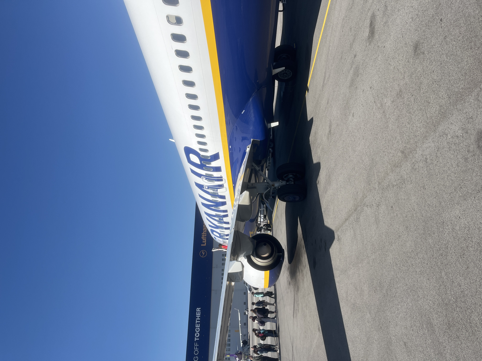 The image shows a Ryanair airplane on the tarmac at Sofia Airport in Sofia, Bulgaria. The plane is blue, white, and yellow. People are boarding or disembarking the aircraft. There is a Lufthansa plane in the background. The sky is blue. The tarmac is gray and has a yellow line.
