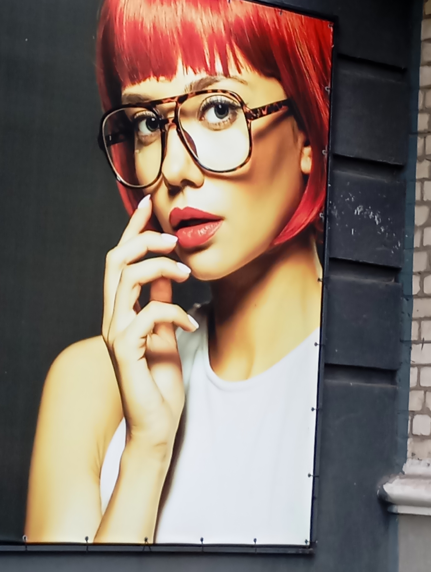 The image is a close-up of a large billboard advertising a woman with bright red hair and large, tortoise-shell glasses. She is wearing a white top and her left hand is raised to her face, touching her lips. The billboard is mounted on a dark, textured wall, likely on the side of a building. The surrounding environment suggests an urban setting, with a brick wall visible on the right side of the frame. The billboard is secured with small metal grommets and clips. Based on available information, this billboard is located in Horishni Plavni, Ukraine.