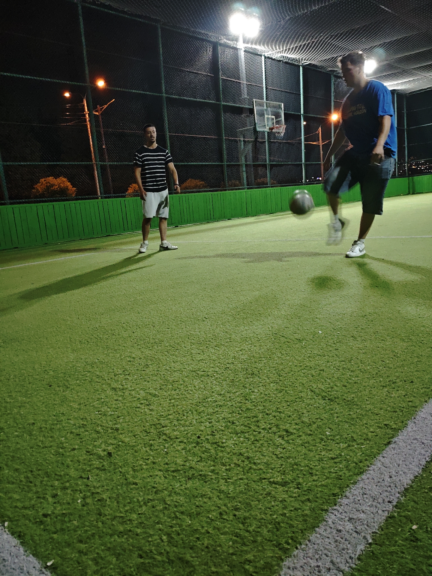 Two men are on an artificial turf soccer field at night. The man on the left is standing and wearing a striped t-shirt and white shorts. The man on the right is in motion, kicking a soccer ball with his left foot. He is wearing a blue t-shirt and dark shorts. A basketball hoop is visible in the background. The field is enclosed by a fence and illuminated by artificial lights. The location is described as Unknown, Unknown.