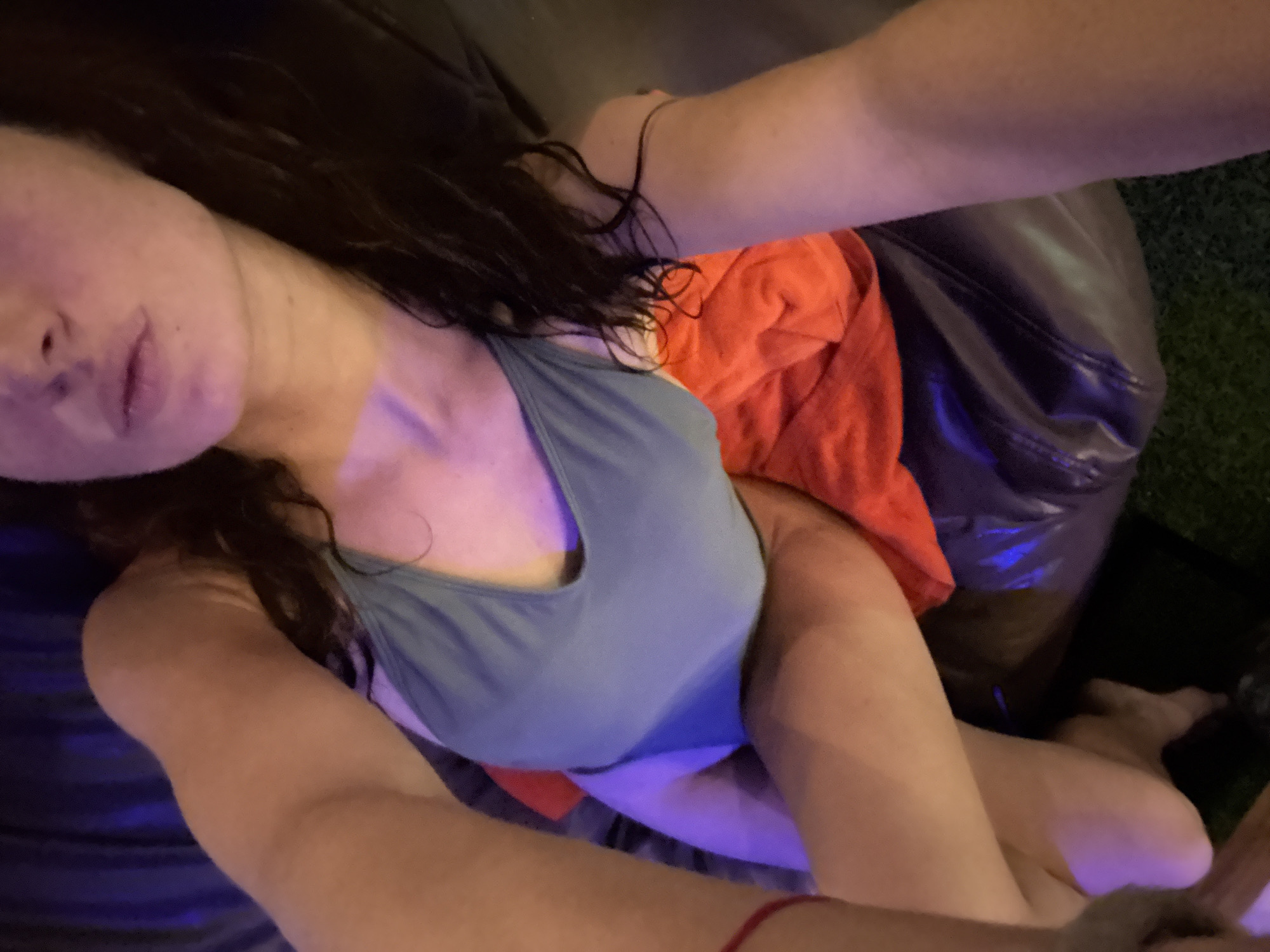 A young woman in a blue swimsuit is positioned on a couch with a black leather surface. She is lying on her side with her legs bent and one arm extended. A red towel is placed beneath her. The background consists of dark blue curtains. The scene is lit with purple and blue lights, creating a dimly lit and atmospheric environment. The location appears to be an indoor setting, possibly a hotel room or a private residence, and is not specified to be in Ko Samui District, Thailand.
