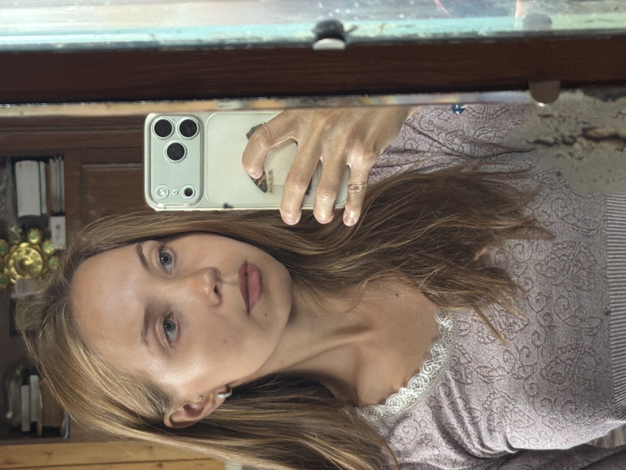 The image is a selfie taken in a mirror, capturing a woman's face and upper body. She has long, light brown hair, blue eyes, and fair skin. She is wearing a light purple shirt with a lace trim. Her left hand is visible, holding a smartphone to take the picture. The phone is in a clear case with a silver camera bump.
In the reflection, shelves filled with books and some decorative items are visible, suggesting an indoor setting, possibly a home or a library. The overall lighting appears to be natural, indicating it might be daytime. The woman's expression is neutral, and the image has a casual, personal feel. The reflection in the mirror shows a wooden frame and a slight distortion, typical of a mirror's surface. The background is slightly out of focus, keeping the attention on the subject. There is no visible text in the image.