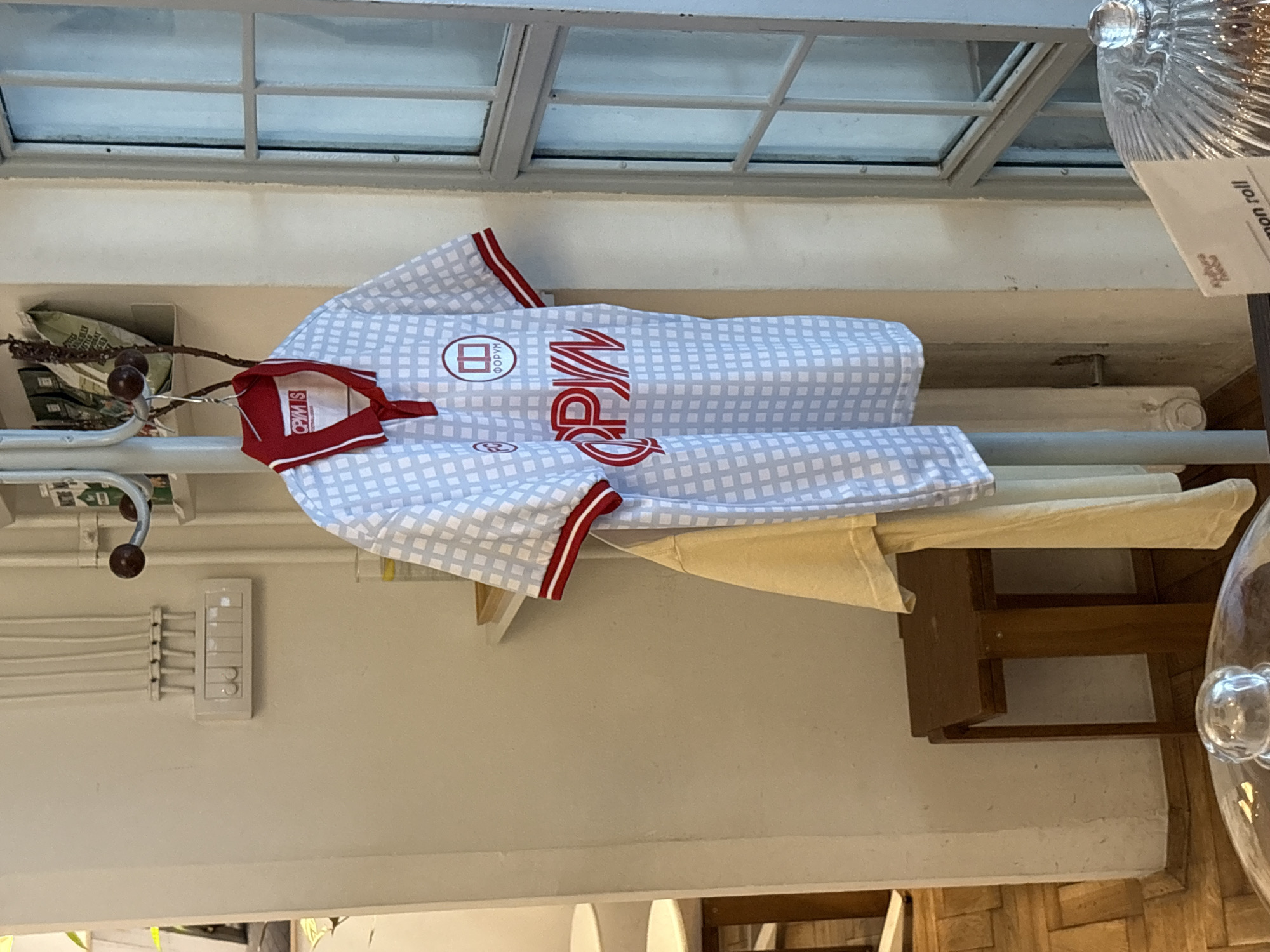 The image shows a light blue and white checkered shirt hanging on a rack, with red trim around the collar and sleeves. The shirt has a logo with Cyrillic text on the left side of the chest and the word "OPYN" in red Cyrillic text on the right. The shirt appears to be a sports jersey, possibly for a Serbian team, given the location context.
The shirt is hanging in what appears to be an indoor setting, possibly a store or a home. Behind the shirt, a rack with some greenery and what looks like packaging is visible. To the right of the shirt, a wooden shelf with a light-colored fabric is also visible. In the background, a window with a grid-like structure is seen, suggesting it's daytime.
A glass object, possibly a vase or a decorative item, is visible on the far right, with a label that appears to be in English, saying "mon roll". A light switch panel is mounted on the wall to the left. The overall impression is a somewhat cluttered but organized display of items. The lighting suggests it's a well-lit indoor space.