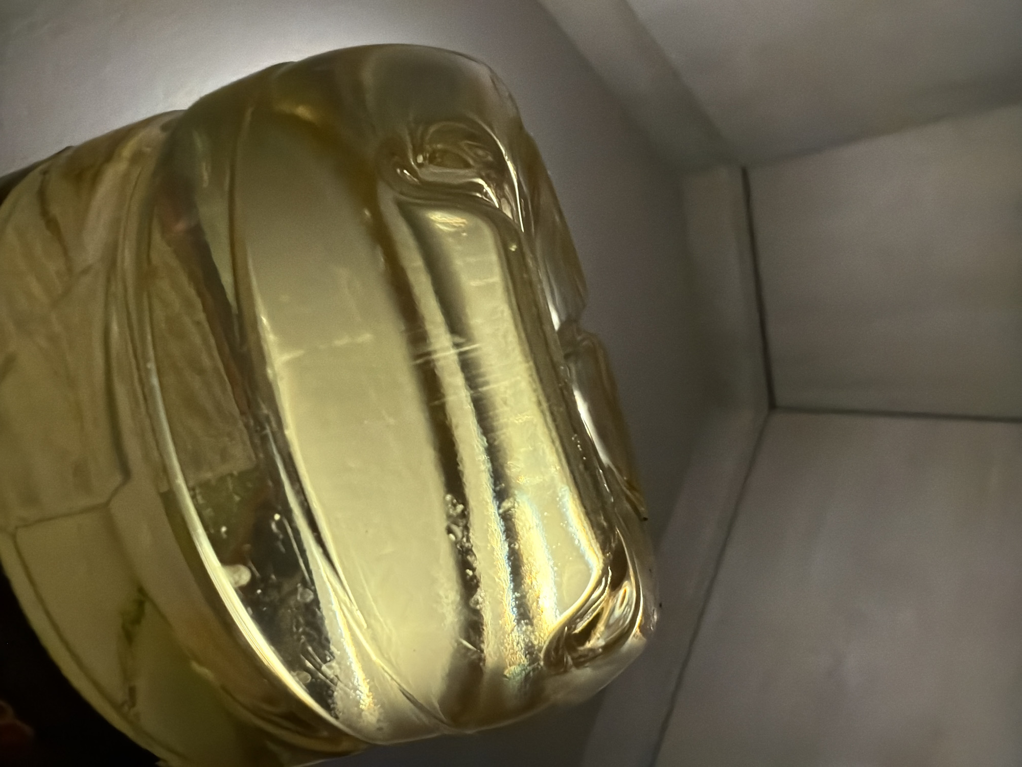 A close-up shot depicts a gold-colored decorative object resembling a human head. The object is made of a transparent, pliable material, possibly plastic or resin, and exhibits distortions and internal imperfections. It appears to be placed in a corner of a room or against a light-colored wall, with the top and side visible. The setting's simple background does not reveal specific clues about the object's location within Budva, Montenegro. There is no evidence of people or interaction with other objects in the frame.