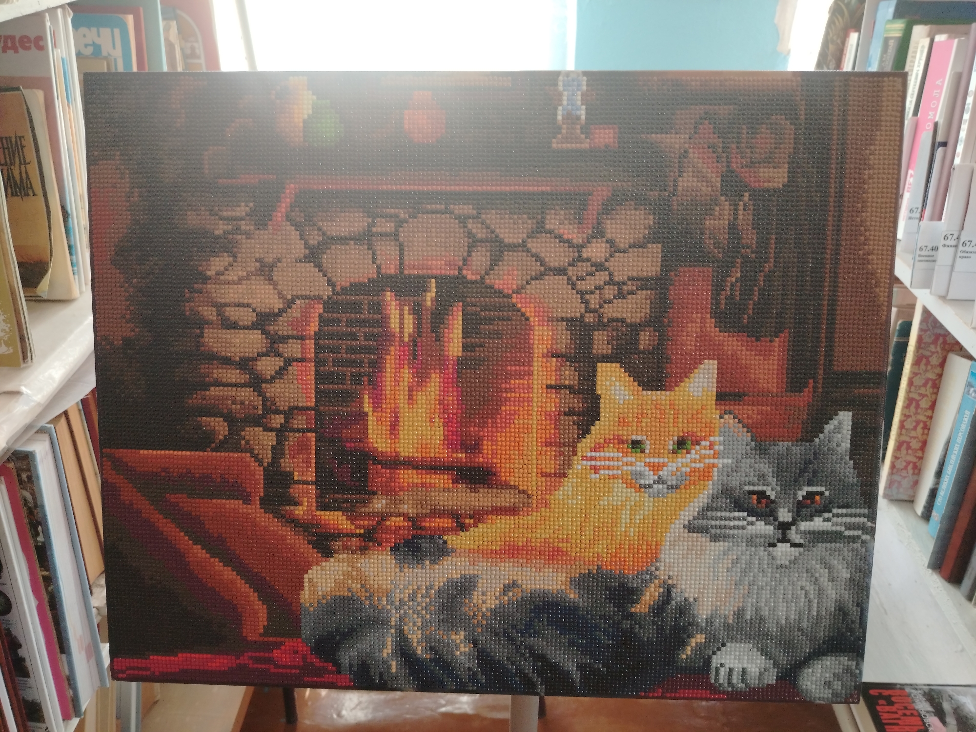 The image shows a diamond painting of two cats sitting in front of a fireplace. The painting is displayed on an easel in what appears to be a library or a room with bookshelves. The painting depicts a warm scene with flames in the fireplace and the cats, one orange and one gray, looking towards the viewer. The background is dark, with hints of furniture and the fireplace structure. The bookshelves in the background are filled with books. The specific location in the city of Klyuchi, Russia, cannot be determined from the visual information provided in the image.