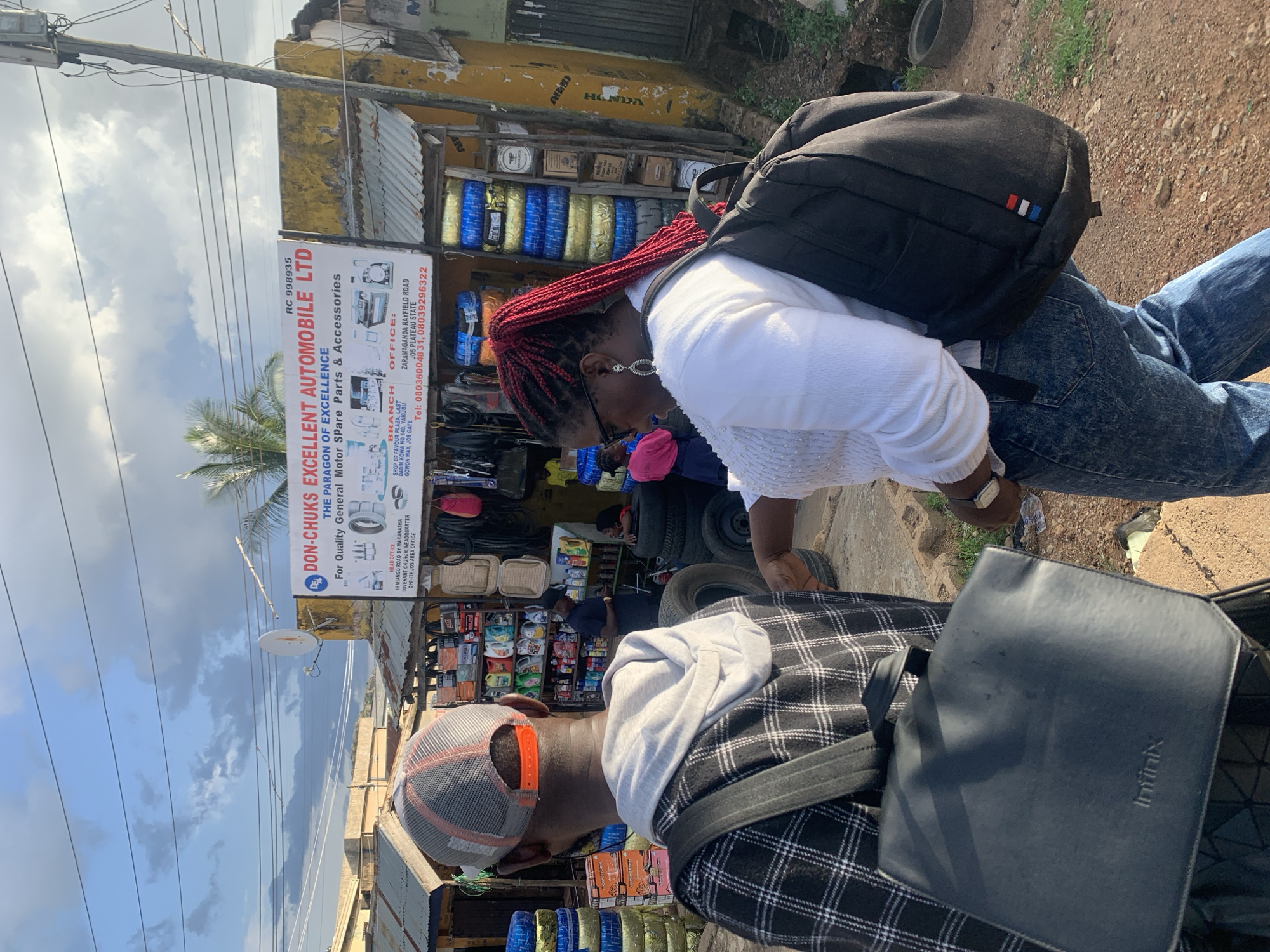 This media file captures a street scene in Jos, Nigeria, featuring two individuals. The primary subject is a woman with vibrant red braided hair, wearing a white long-sleeved top and blue jeans, with a black backpack slung over her shoulders. She appears to be looking down or to the side, with a slight bend in her posture, suggesting engagement with something or someone not fully in frame. She is accessorized with hoop earrings and a watch on her left wrist.
Beside her, a person wearing a checkered shirt over a hoodie and a grey baseball cap is visible from the side, their face mostly obscured by the cap. A dark grey bag with a geometric pattern is draped across their lap.
The background reveals a bustling outdoor market or shop setting. A prominent sign reads "DON-CHUKS EXCELLENT AUTOMOBILE LTD" with details about their "General Motor Spare Parts & Accessories". Shelves stocked with tires, car parts, and various other goods are visible, indicating a commercial area. The architecture appears modest, with a yellow-painted building and corrugated metal roofing. The ground is unpaved, composed of gravel and dirt. The sky is visible above, with clouds suggesting it might be daytime, possibly late afternoon given the lighting. The overall atmosphere seems casual and everyday. The presence of the shop sign and the goods displayed strongly suggests a commercial environment focused on vehicle parts and accessories. The distant view shows more of the urban landscape.
