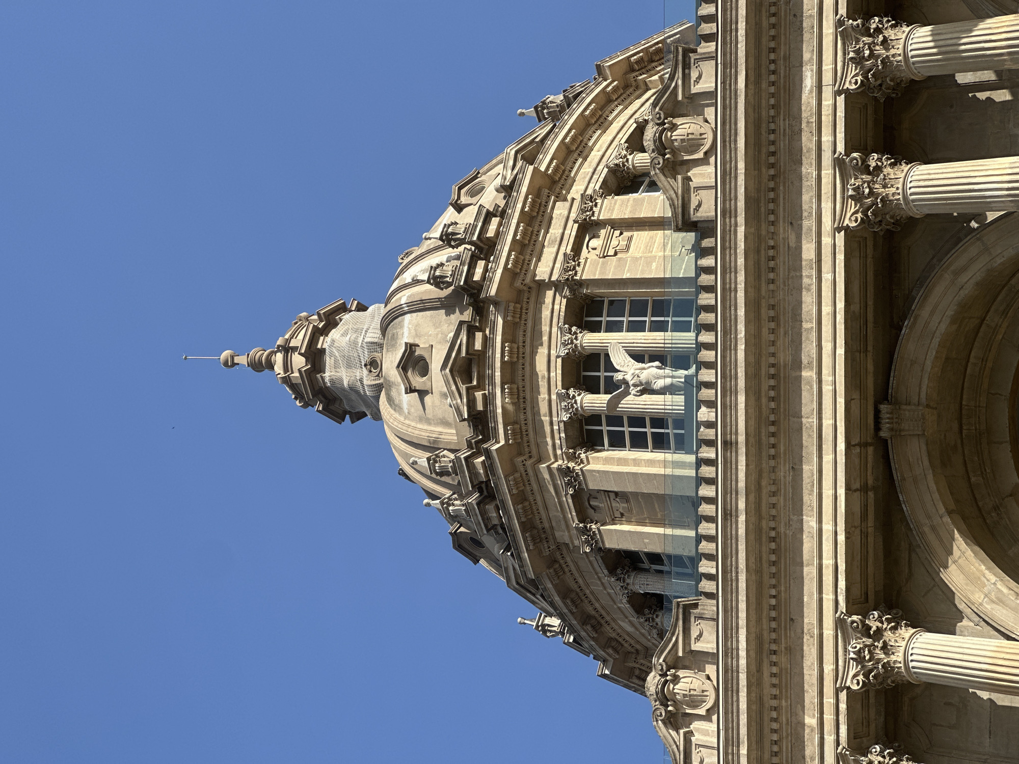 The image shows the dome of the National Palace of Montjuïc in Barcelona, Spain, set against a clear blue sky. The dome is ornate, made of stone, and features intricate architectural details such as columns, carvings, and a central spire. A white statue of an angel is visible between two windows on the building's facade. Below the dome, the building's lower levels display more classical architectural elements, including large fluted columns and arched entrances. A modern glass barrier runs along a balcony or terrace. The lighting suggests it is daytime.