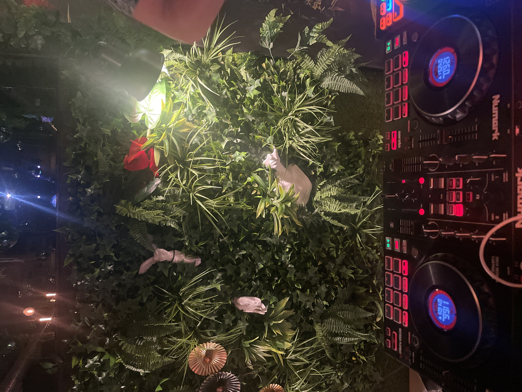 The image depicts a vertical arrangement of artificial greenery with interspersed faux flowers and ornamental fans. To the right, a Numark DJ controller is partially visible, its illuminated buttons and dials suggesting active use. A spotlight is aimed at the greenery, creating highlights and shadows. The overall composition suggests a decorative or event setting. The location cannot be determined from the image, so Sofia, Bulgaria is unknown.
