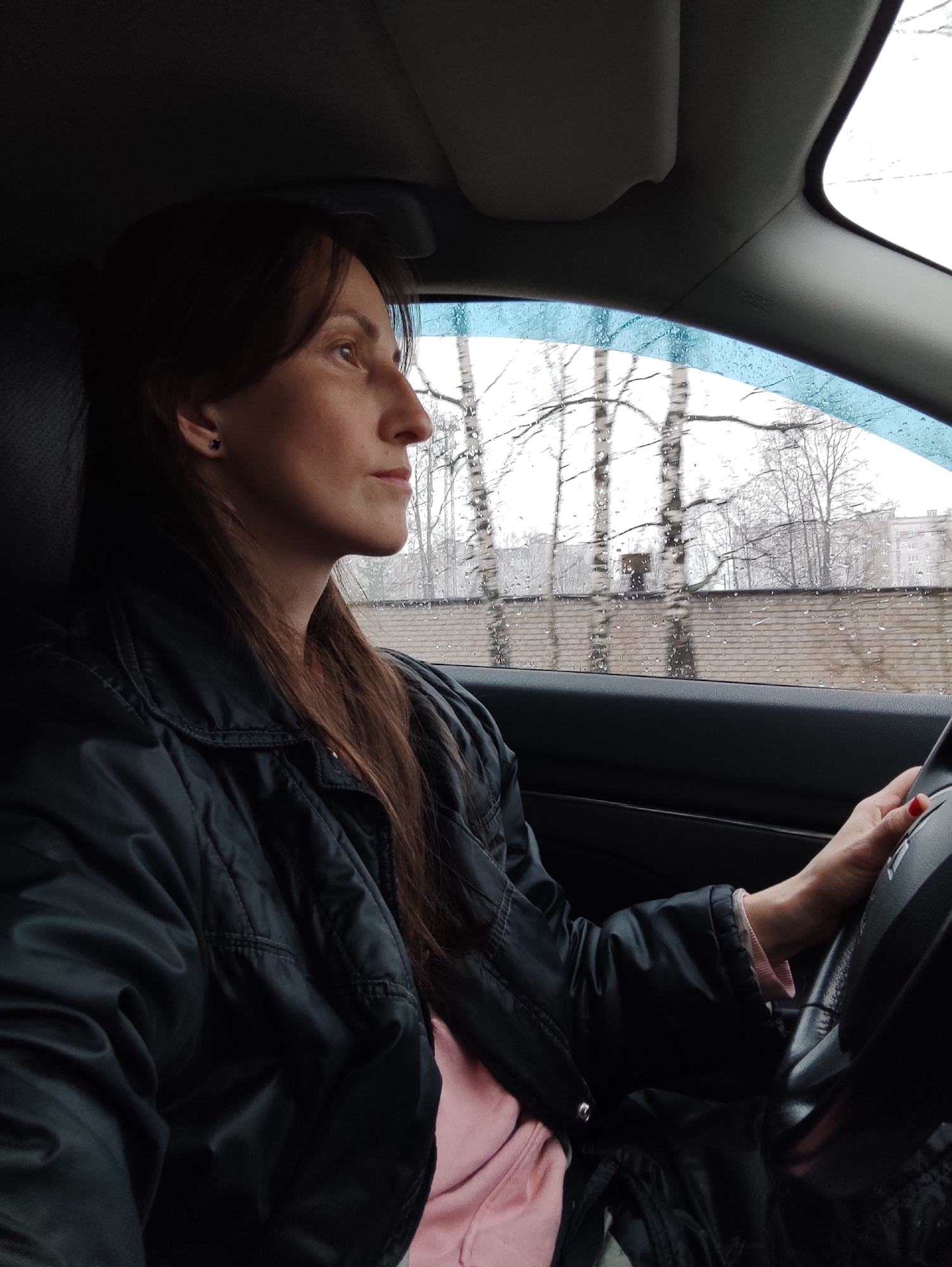 The image shows a woman driving a car. It appears to be a rainy day, as there are raindrops on the car window. The woman is wearing a black jacket and a light pink shirt. She has long brown hair. Outside the car window are birch trees. The location appears to be in Chaikovsky, Russia, based on the architecture of the buildings visible in the background.