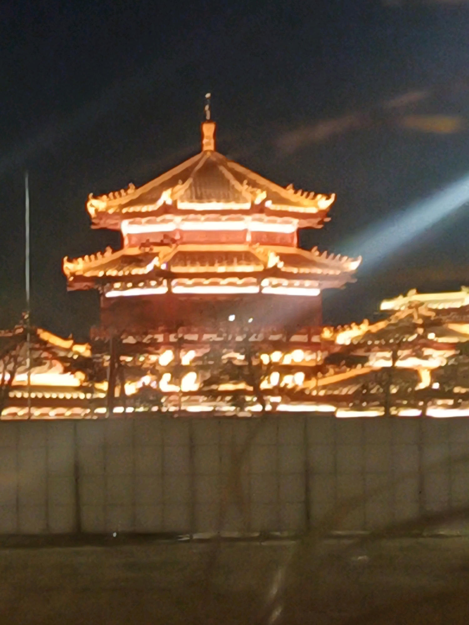 The image shows a traditional East Asian-style building illuminated by orange lights at night. The building has multiple tiered roofs with upturned eaves. In the foreground, there is a light-colored wall or barrier, and beyond that, a dark ground surface. The sky is dark, with some faint clouds visible and a bright light beam cutting across the upper right portion of the image. There are no people visible in the image. This structure is the Lumina Tower at PIK 2 City, North Jakarta, Indonesia.