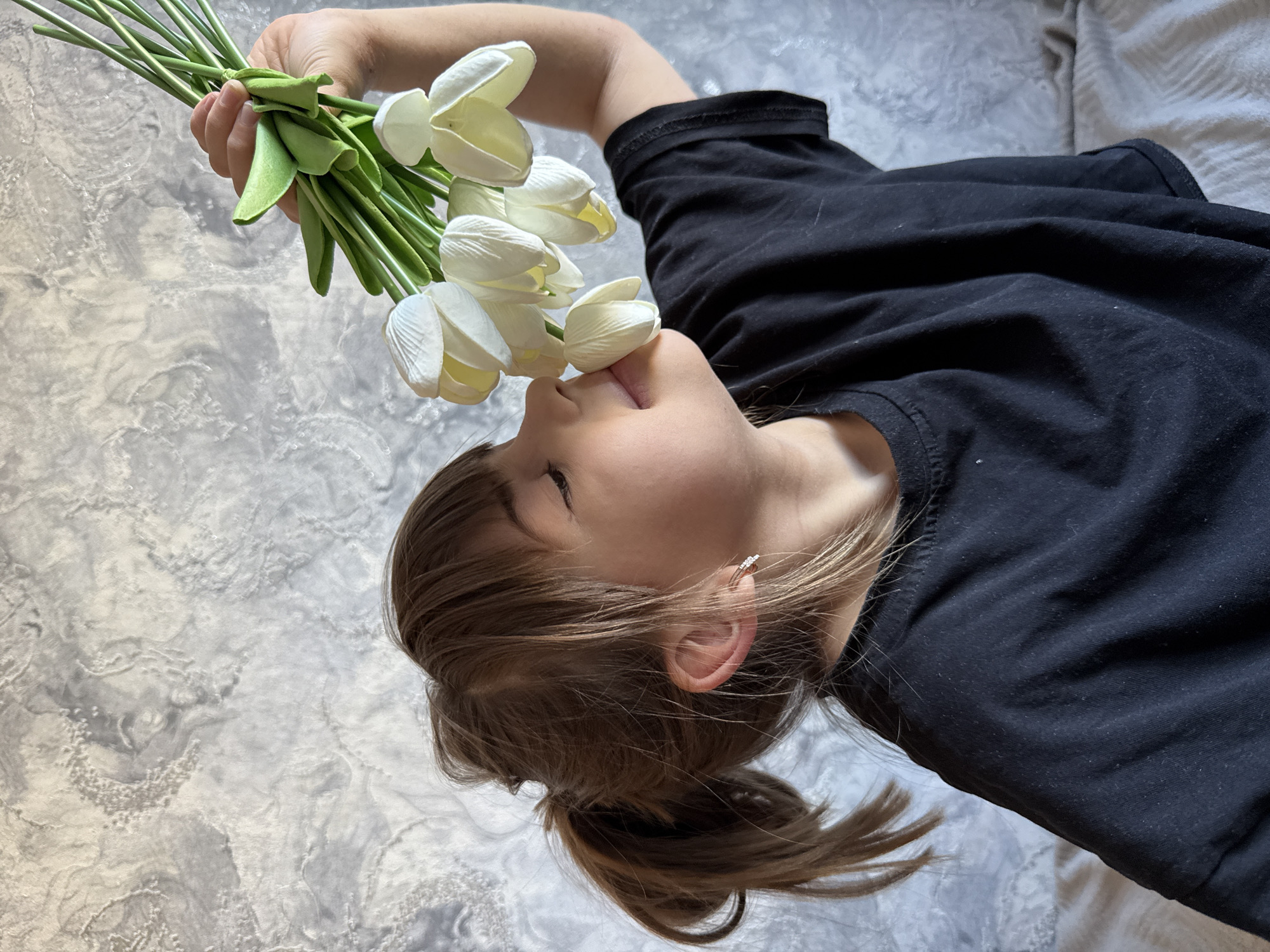 A girl lies on her back on a gray textured surface, possibly a bed, in an unknown location. She wears a black t-shirt and has her light brown hair pulled back into a ponytail. Her right ear is visible, adorned with a small, circular earring. In her right hand, she holds a bouquet of white tulips, the flowers positioned close to her mouth, with her tongue slightly extended towards them. The bouquet consists of several stems bound together with a green tie. The background is a mottled gray.