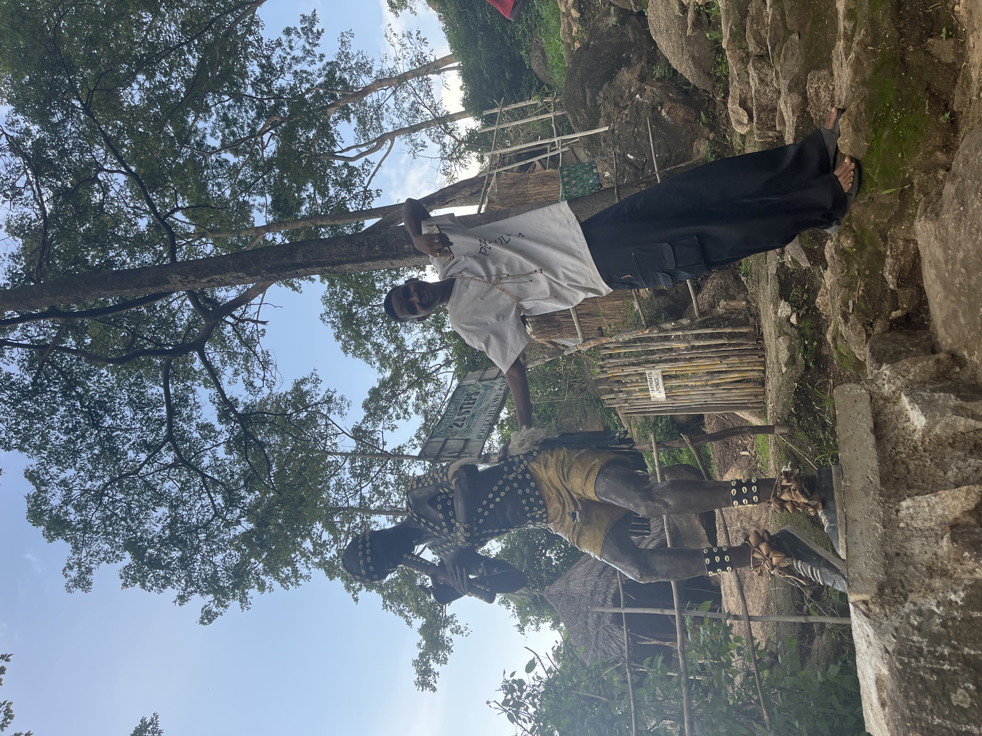 In Jos, Nigeria, a young man stands to the right of a life-sized statue of a person playing a flute. The statue, clad in traditional attire with shells and a headdress, is positioned on a stone pedestal. The man, wearing a white t-shirt and black cargo pants, smiles and poses with his left arm extended towards the statue, his right hand on his hip. Behind them, a sign reads "Ogechukwu Park" and mentions "25 Steps." The background features lush green trees, a thatched-roof hut, and a rocky terrain, indicative of an outdoor tourist attraction. The sky above is a clear blue with scattered clouds.