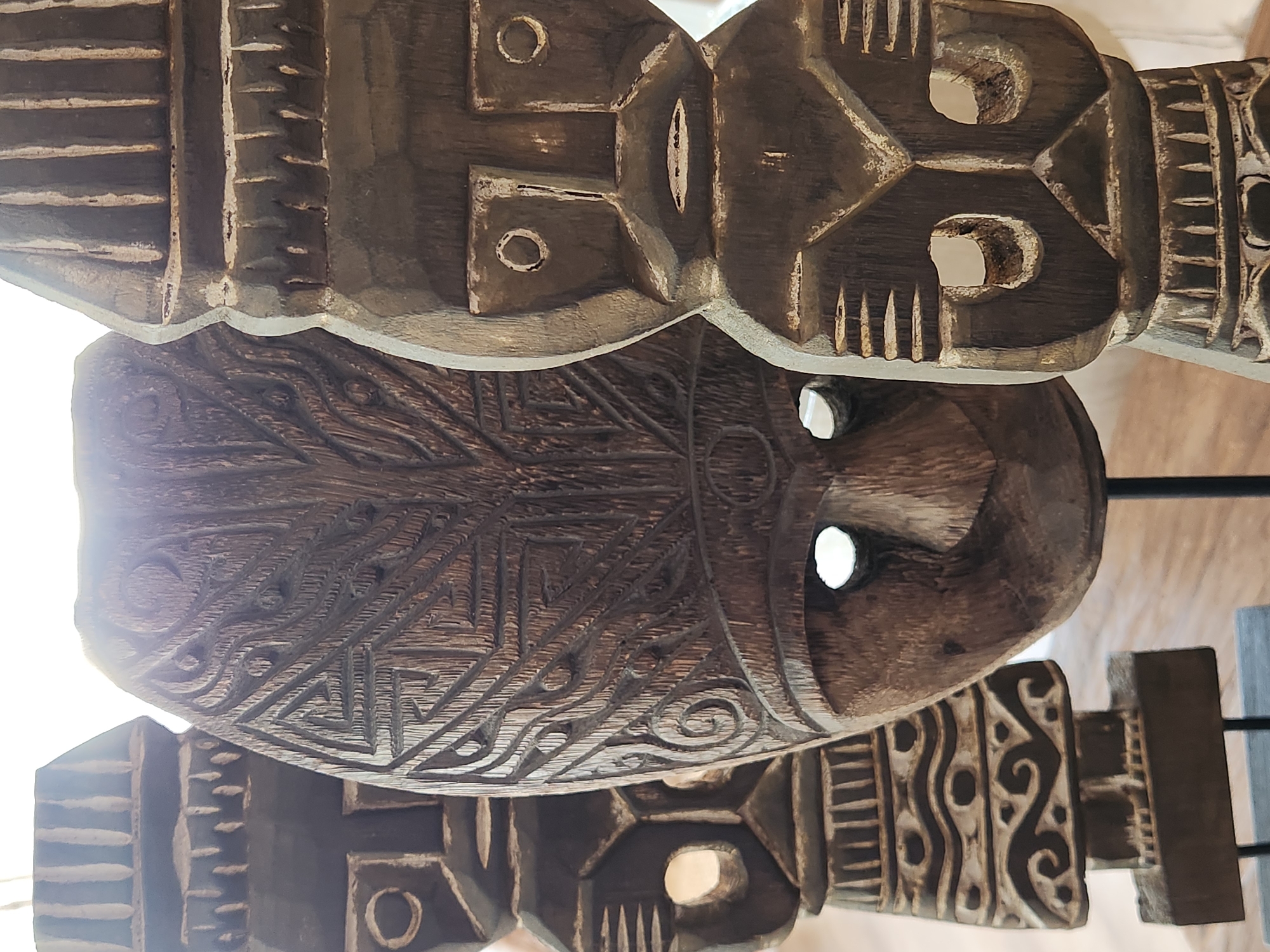 The image shows a close-up, vertically oriented view of several intricately carved wooden masks. The masks appear to be ancient or tribal in origin, with one prominently displayed in the foreground. This central mask is oval-shaped and features detailed geometric patterns and stylized facial features, including two eye openings. The wood has a rich, dark brown patina, and some areas are highlighted with a lighter, possibly white or light gray, accent color, suggesting age or a deliberate decorative technique.
Behind the central mask, parts of other masks are visible, each with distinct carving styles. One mask above the central one has a more abstract, angular design with circular and elongated openings for eyes. Another mask below the central one exhibits a different pattern of parallel lines and geometric shapes. The texture of the wood is evident, showing the marks of carving tools.
The lighting in the image is strong and suggests it might be daytime, possibly outdoors or in a well-lit indoor space. The bright light causes some areas of the masks to be overexposed, particularly on the left side, and creates highlights and shadows that emphasize the depth and detail of the carvings.
The overall impression is one of artistry and cultural heritage. The masks are arranged in a way that suggests they are either part of a collection, displayed for exhibition, or perhaps part of a larger decorative installation. There is no visible text within the image. The setting appears to be in Maitland, United States of America, but specific location cues within the image are absent.