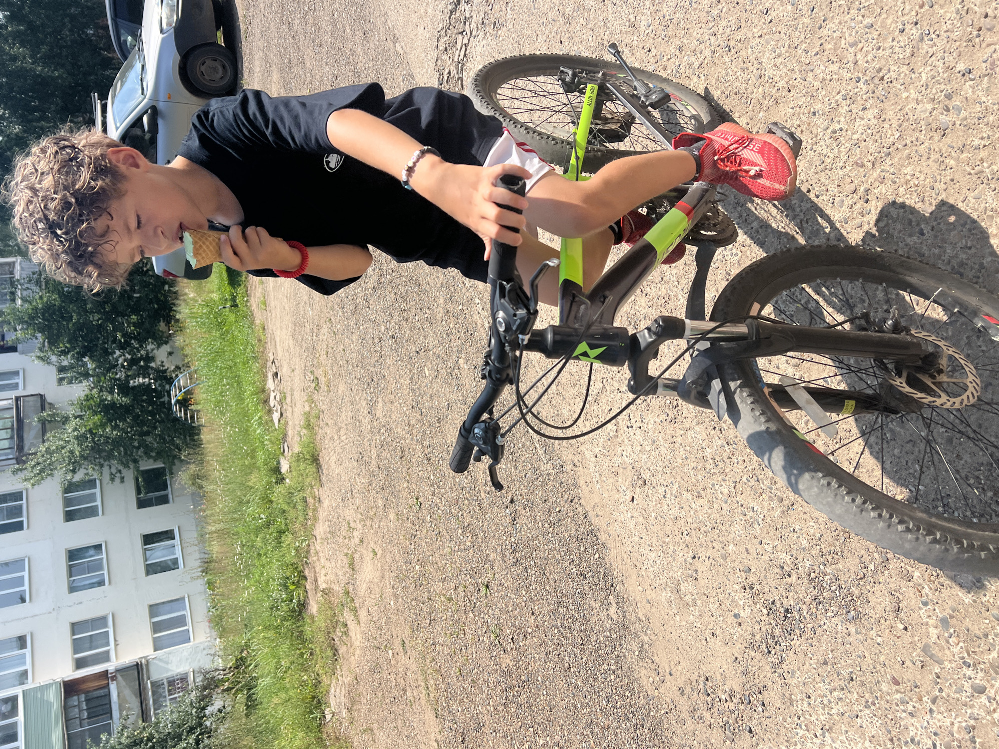 A young boy with curly brown hair is riding a bicycle and eating an ice cream cone outdoors. The boy is wearing a black t-shirt, white shorts with red trim, and red sneakers. He has a red bracelet on his left wrist and a beaded bracelet on his right wrist. The bicycle is a dark grey and green mountain bike. The scene appears to be in a residential area, with a multi-story building in the background and a car parked nearby. The ground is covered in gravel and dirt. The overall environment is sunny and appears to be a warm day.