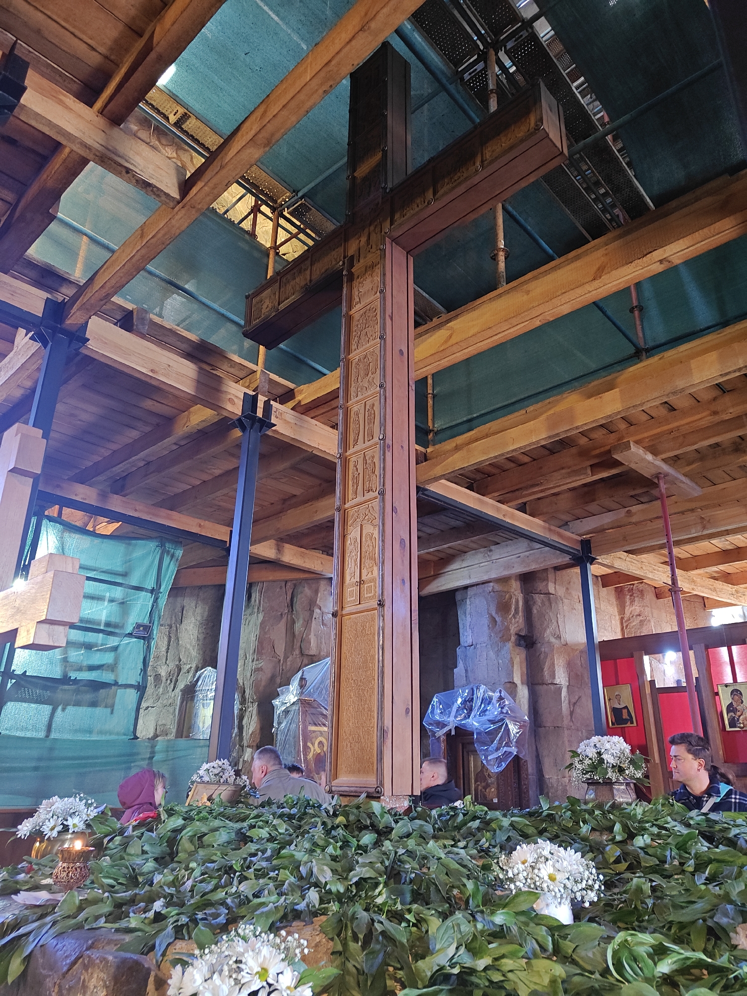 The image shows the interior of a building in Unknown, Unknown. A large wooden cross dominates the scene, extending from the ground to the ceiling. The cross is adorned with carved panels. The ceiling is constructed of wood beams and metal supports. Scaffolding and green mesh are visible above the beams.
In the foreground, there is a display of greenery and white flowers, partially obscuring several people. A woman with a red head covering and a man in a green jacket can be seen on the left. Further to the right, two more individuals are visible, including a man in a plaid shirt. There are candles on the left and religious images on the right. The walls are made of rough-hewn stone.
