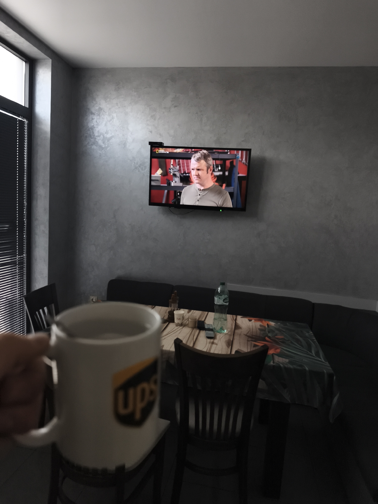 A person's hand holds a white mug with the UPS logo in the foreground. The mug contains a light-colored liquid. In the background, a television screen displays a man wearing a grey shirt. The television is mounted on a textured grey wall. A dining table with a patterned tablecloth is visible below the TV, with chairs around it. A bottle of water and some other objects are on the table. The scene appears to be indoors, likely a dining or living area. The lighting suggests it might be daytime, but the interior lighting is dim.