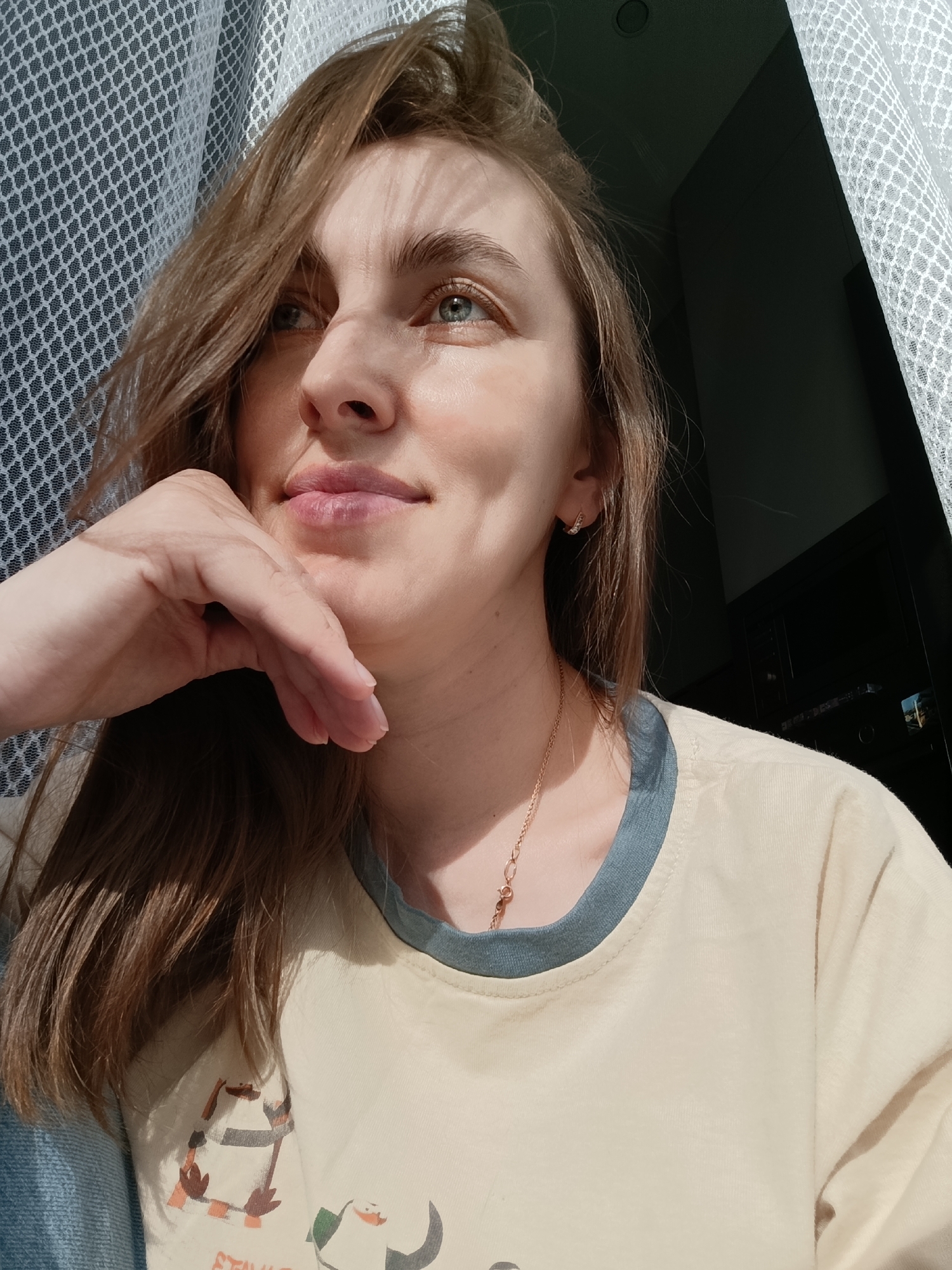 A woman is captured in a close-up shot, her face partially illuminated by sunlight filtering through a white, net-like curtain. Her hand is positioned beneath her chin in a pensive pose. She is gazing upwards and slightly to the left, exhibiting a subtle smile. Her hair is brown and shoulder-length, with some strands falling across her face. She is wearing a light-yellow t-shirt with a blue trim around the neck and a delicate gold necklace. One earring is visible. The location appears to be indoors, with a dark, undefined area behind her suggesting a room or window. The exact location in Chaikovsky, Russia cannot be determined from the image alone.