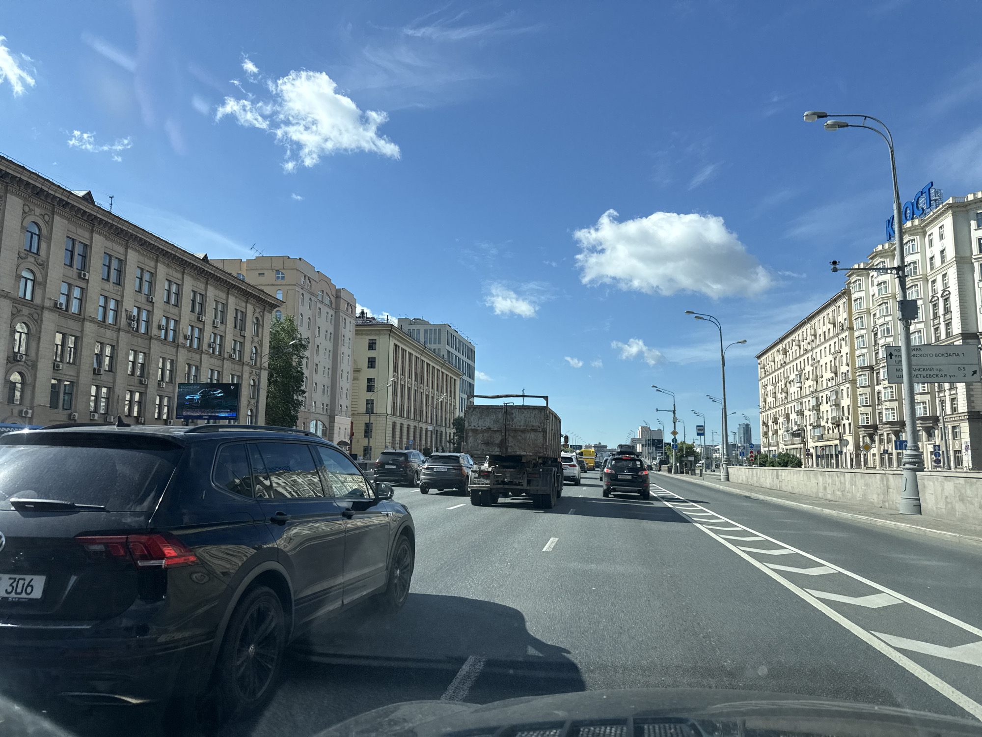 The image depicts a street scene in Moscow, Russia, on a sunny day with a bright blue sky and scattered white clouds. The perspective is from inside a vehicle, looking forward at traffic. On the left, an apartment building with many windows and air conditioning units lines the street. To the right, a similar, longer apartment building extends into the distance. The street itself is a multi-lane road with visible lane markings and traffic. Several cars and a large truck are on the road, some moving and some stationary. A black SUV with license plate "306" is prominently visible in the foreground on the left. In the distance, more buildings and streetlights are visible, along with signage, some of which appears to be in Cyrillic script. The overall impression is of a busy urban environment during daylight hours.