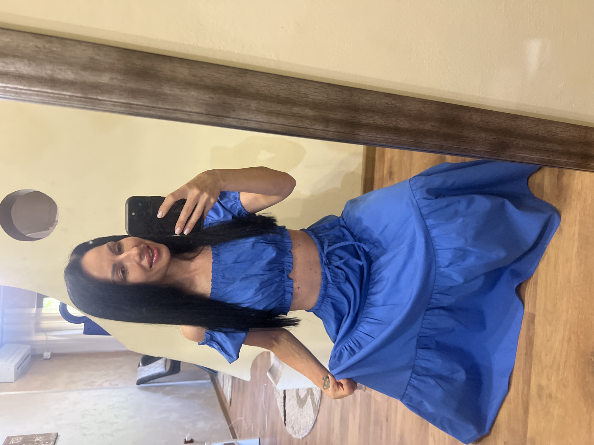 A woman is taking a mirror selfie in a room with light yellow walls and a wooden floor. She is wearing a blue off-the-shoulder crop top and a matching blue tiered maxi skirt. Her dark hair is long and straight. She is kneeling on the floor, holding up the skirt with her left hand and her phone with her right. The room appears to be a residential interior.