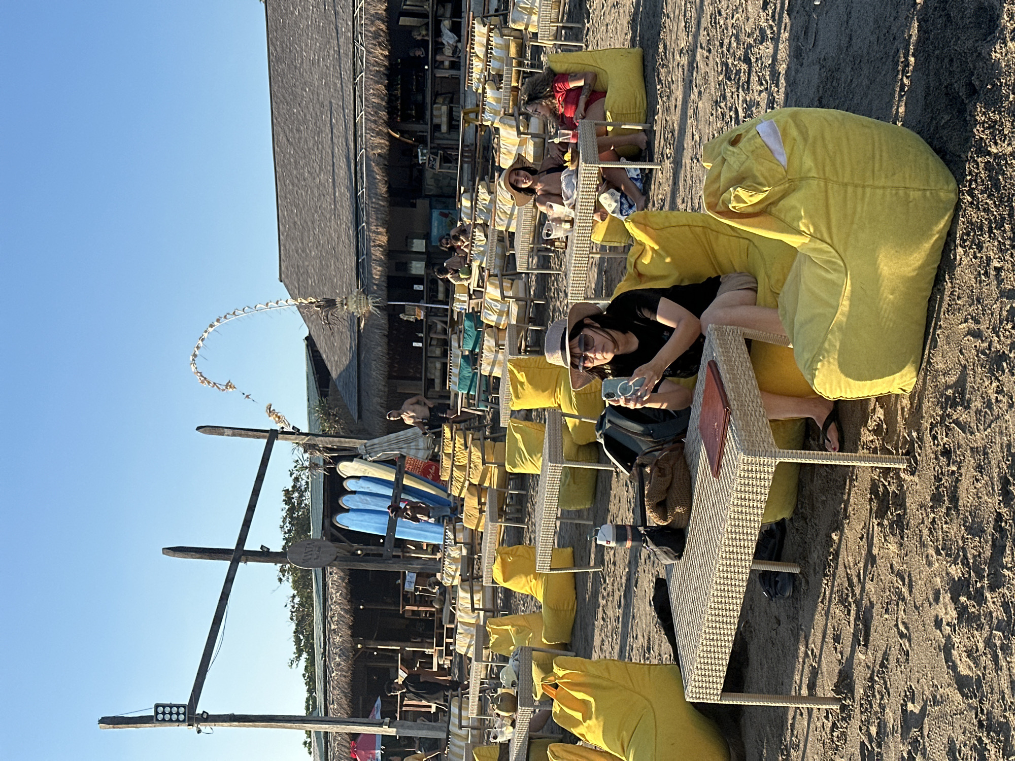 The image depicts a beachside seating area in North Kuta, Indonesia. The scene is set on a sandy beach with a clear blue sky overhead. In the foreground, several yellow beanbag chairs and wicker tables are arranged. One woman, wearing a hat and sunglasses, is seated at a table, looking at her phone. She is dressed in a black t-shirt and shorts. Next to her is a beanbag chair with a cartoon-like face.
Further back, there are rows of yellow and white striped seating with tables, some occupied by people. Behind the seating area is a rustic building with a thatched roof, likely a restaurant or bar. Surfboards are propped up against a wooden structure in front of the building. Various people are visible in the background, some standing and others seated. The overall atmosphere suggests a relaxed, outdoor dining or lounging environment on a sunny day.