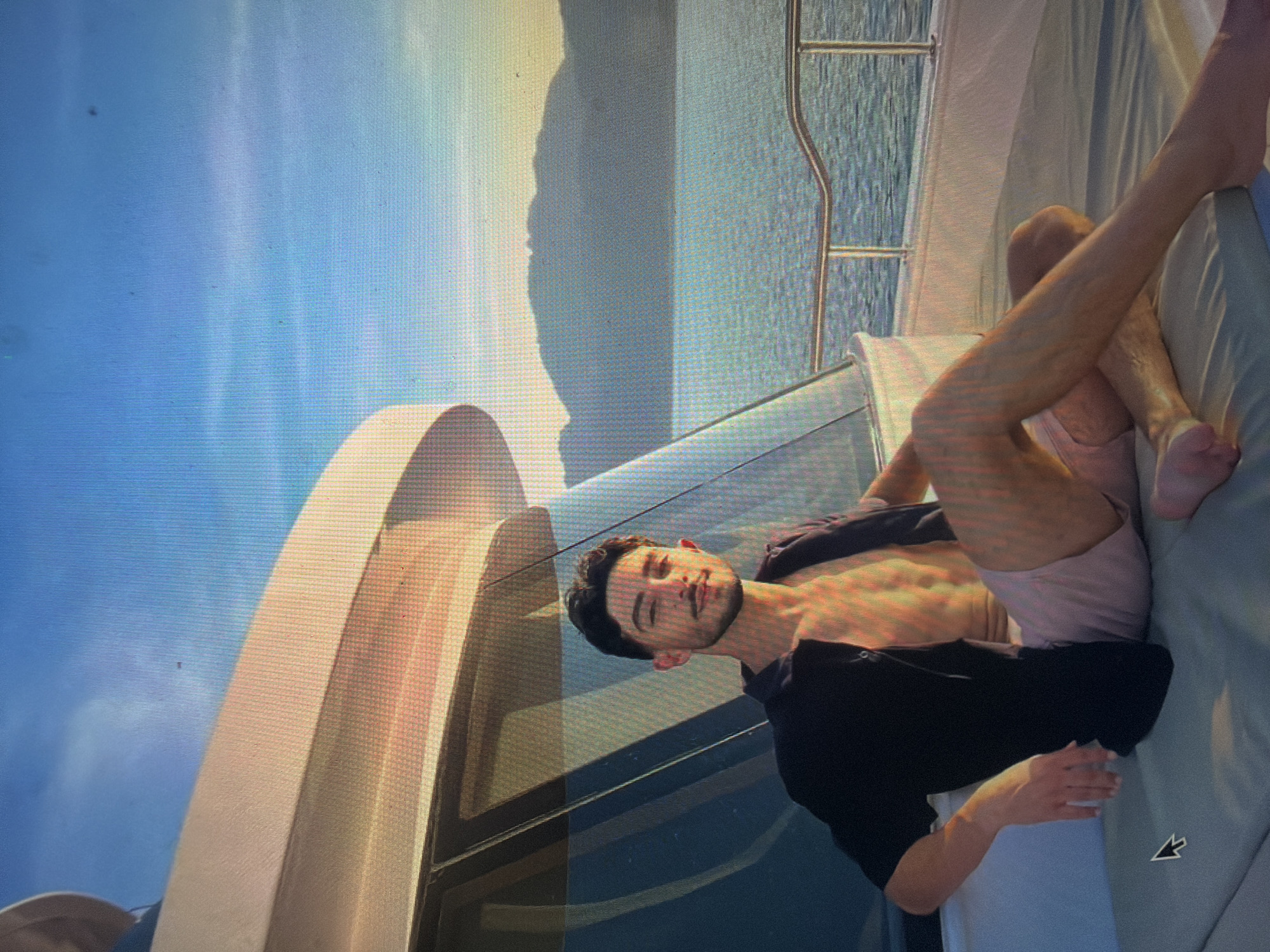A young adult male is seated in a relaxed pose on the cushioned deck of a yacht, observed in a photograph taken of a digital screen. He is light-skinned with dark hair and a short beard and mustache, looking towards the viewer with a slight smile. He wears an open dark short-sleeved shirt, revealing his bare torso, and light pink shorts. His left arm rests on a white armrest, while his right arm is partially obscured behind him. His legs are extended across the cushioned surface.
The yacht features white hull structures, tinted glass windows, and metal railings visible in the background on the right. The setting sun casts warm, golden light across the yacht's surfaces and the man's skin, indicating either late afternoon or early morning.
Beyond the yacht, a vast body of water, likely a sea or large lake, extends to the horizon. Distant, dark-silhouetted mountains line the far shore. The sky above is clear, transitioning from deep blue overhead to lighter, warmer tones near the horizon.
A black computer mouse cursor is visible in the lower-left corner of the image, indicating the image is a capture of a digital display. The image does not contain any discernible landmarks or specific details to confirm the location as Shumen, Bulgaria, or any other specific city.