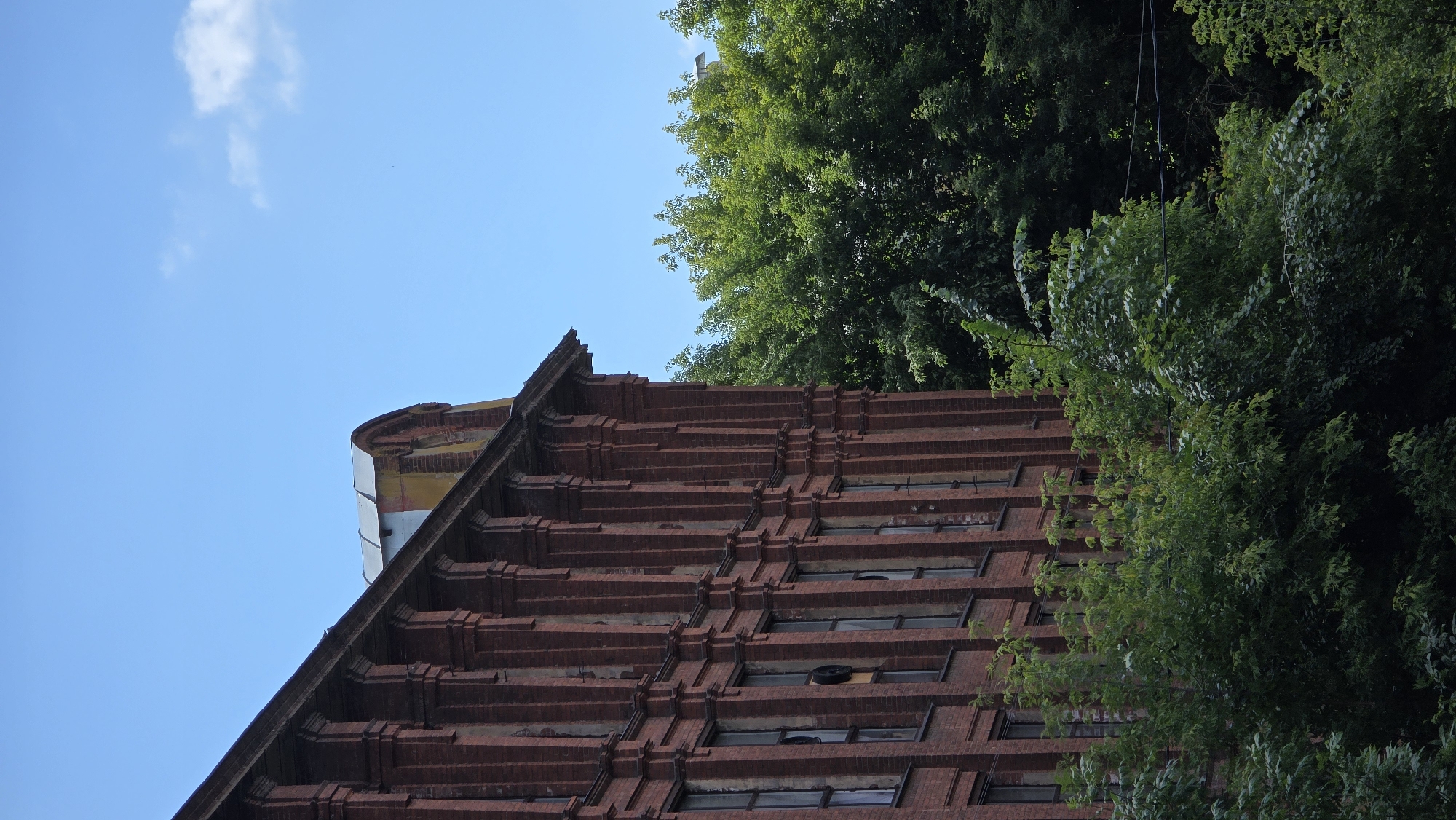 The image shows the side of an old, multi-story brick building with a series of arched windows. The brickwork is a deep red, and the building appears to have a decorative facade with pilasters. The roofline is visible, with a curved, metal-covered section at the top, possibly an attic dormer. The building is partially obscured by lush green trees and foliage in the foreground and to the right. The sky is clear and blue with a few scattered clouds. This building is located in the city of Ivanteyevka, Russia. There are no people or animals visible in the image, and no specific actions are depicted.