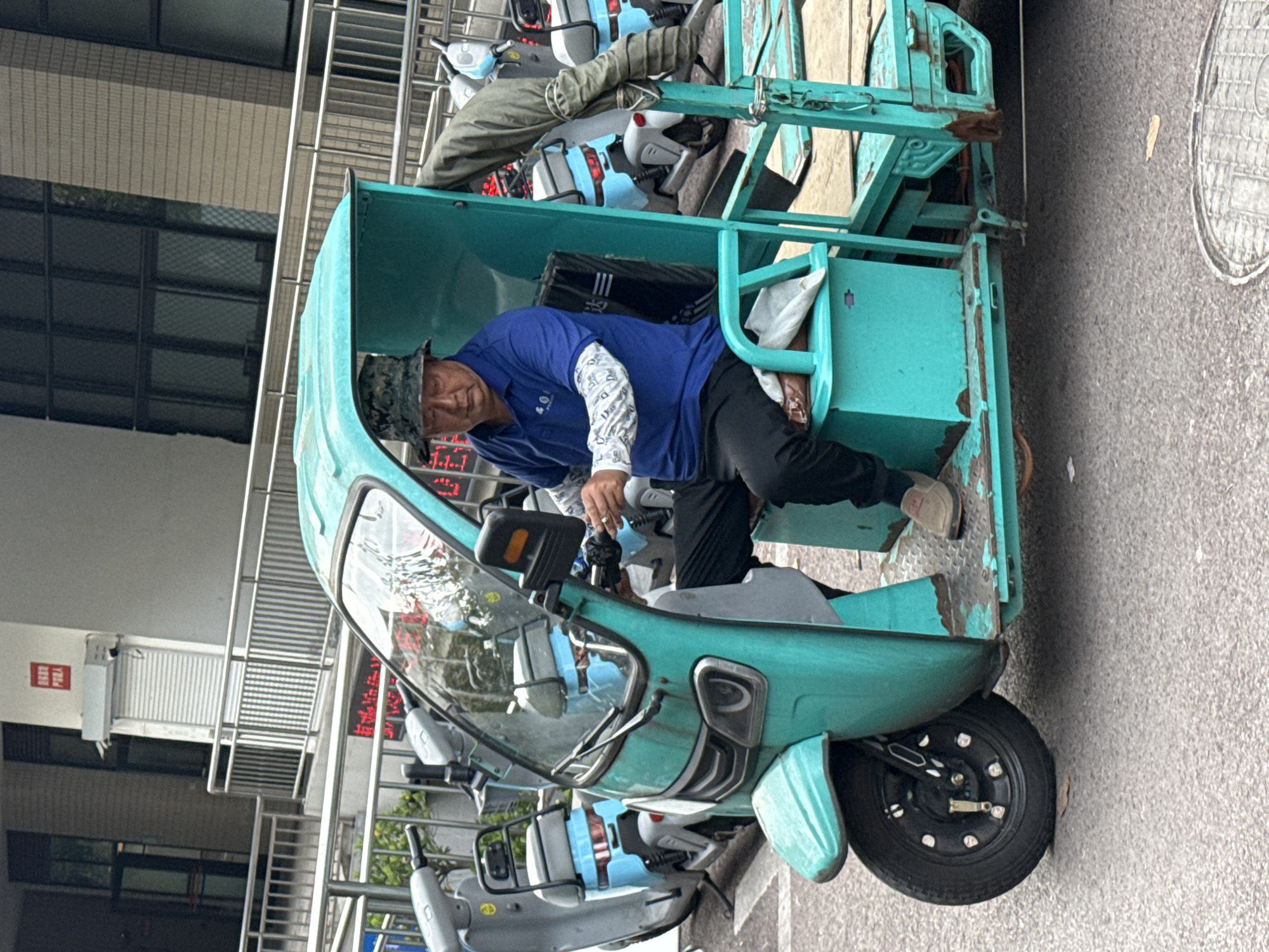 The image shows an elderly Asian man in Jiangjin, China, sitting in a turquoise three-wheeled vehicle. He wears a blue shirt with patterned sleeves, black pants, and a camouflage bucket hat. He is positioned behind the handlebars, appearing to be operating the vehicle. The vehicle is parked on a street next to a building with metal railings and Chinese text on the side. Behind the man's vehicle are other parked motorized scooters or bicycles. The setting seems to be an urban area.