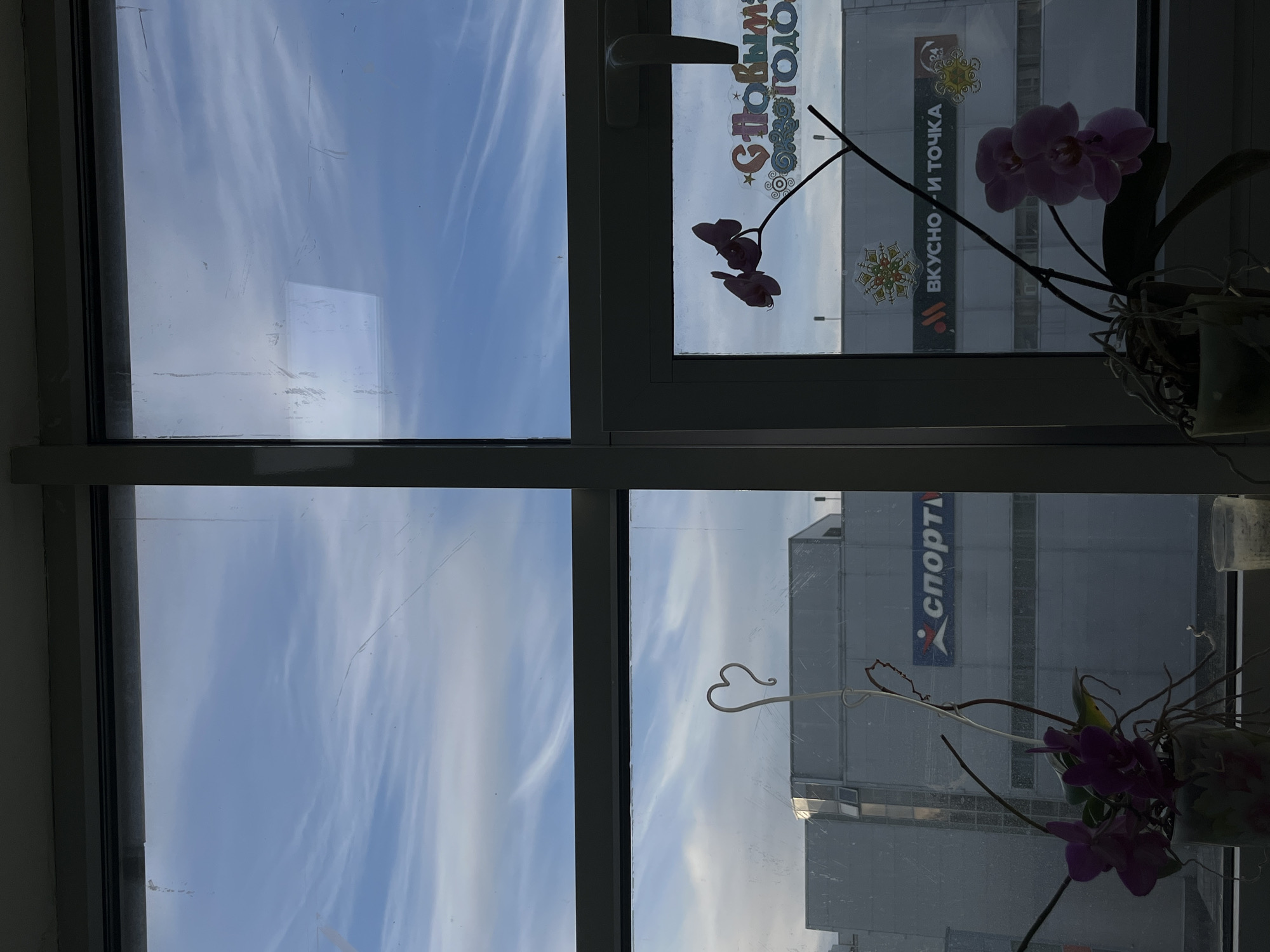 A window view from an interior location in Klin, Russia, reveals a four-paned window, with the top right pane featuring the phrase "С новым годом" in Cyrillic, which translates to "Happy New Year" along with a banner that says "Вкусно и точка". The lower right pane shows a building with the word "СПОРТМ" printed vertically on its facade, adjacent to an orchid plant in a pot, positioned close to the window. The upper left and lower left panes present a view of the sky with thin, streaky clouds. The view through the windows reflects the urban environment of Klin.